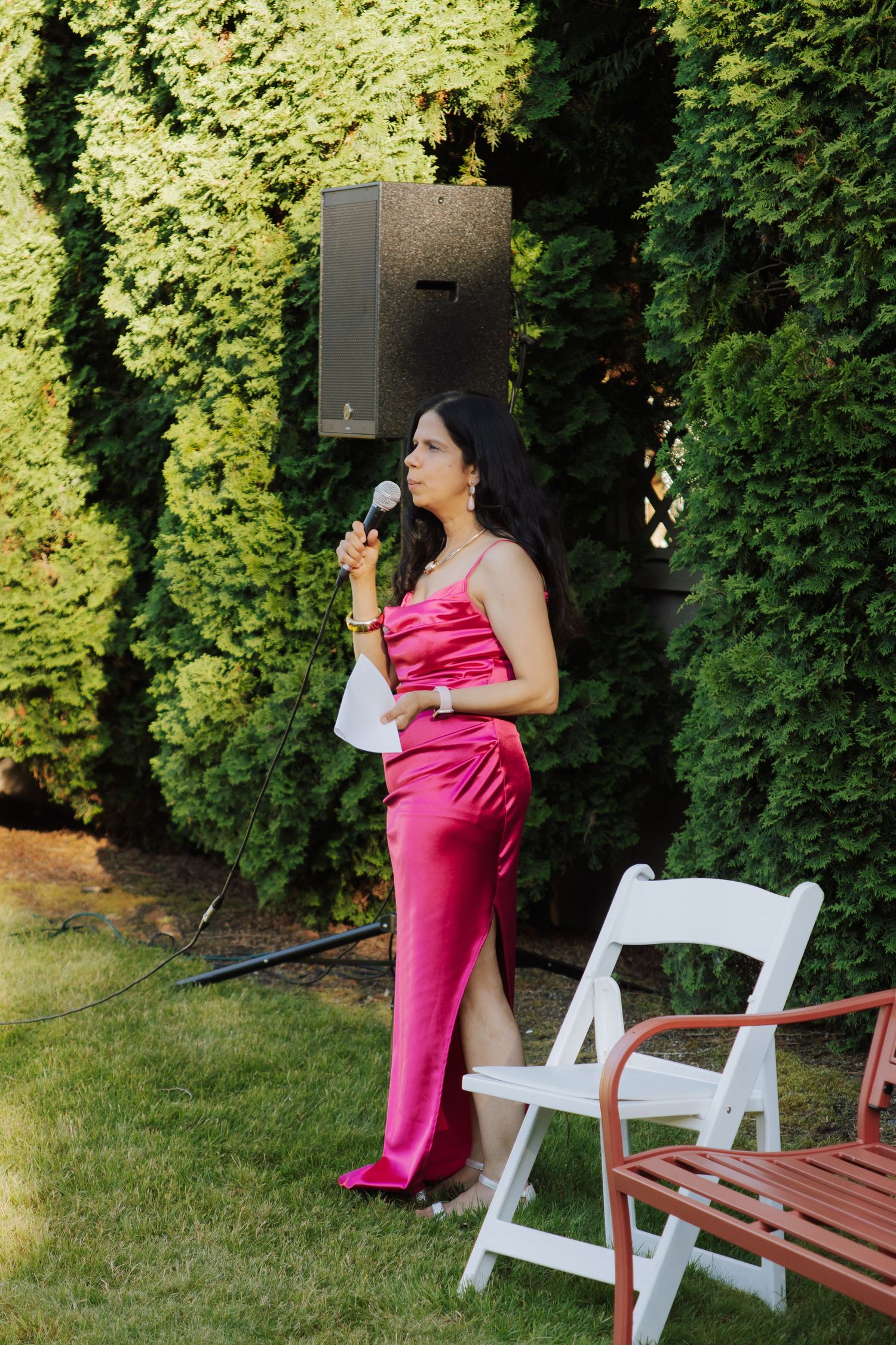 Woman in pink satin gown speaking into microphone at outdoor garden party event.