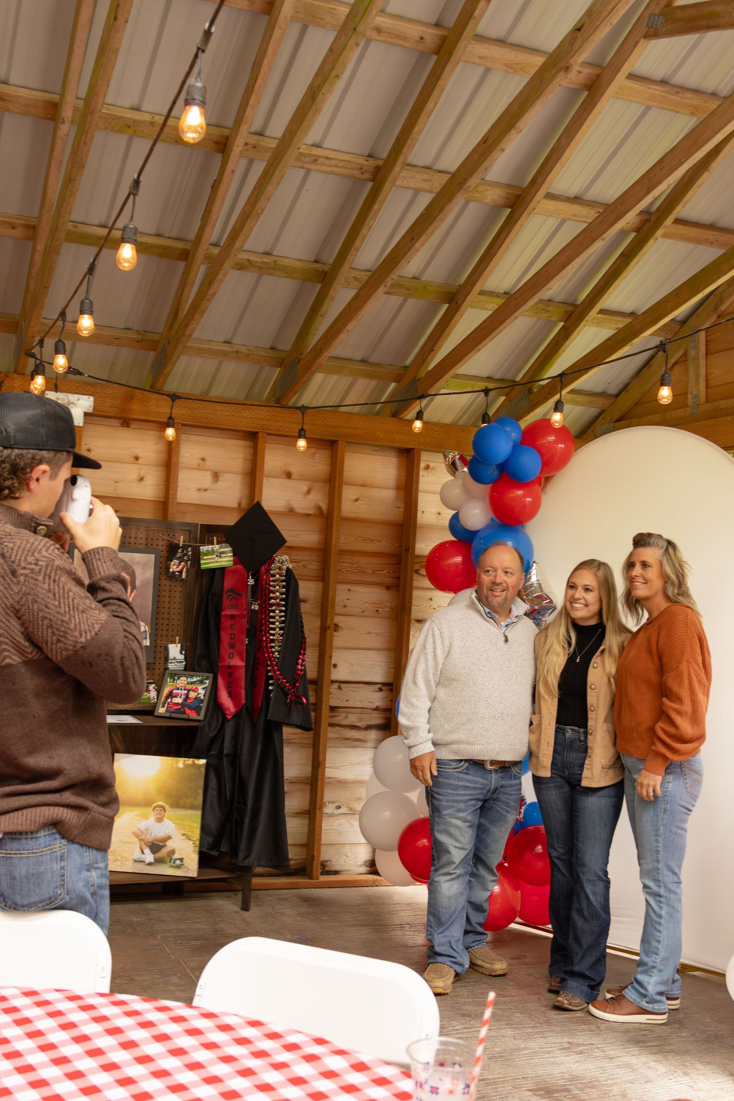 Man photographing couple posing near red white blue balloon arch inside rustic barn.