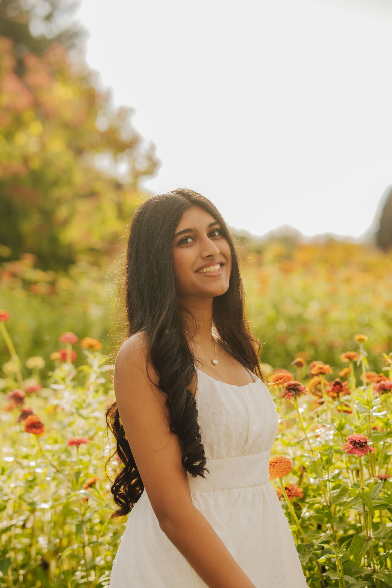 Smiling young woman with long wavy dark hair in a white sundress stands in a sunlit field of orange and pink zinnia flowers.