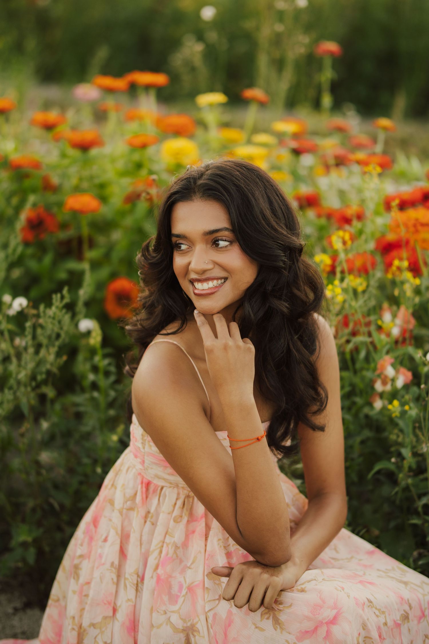 Smiling woman with long wavy dark hair in a floral sundress poses among blooming orange and yellow zinnias in a sunlit flower field.