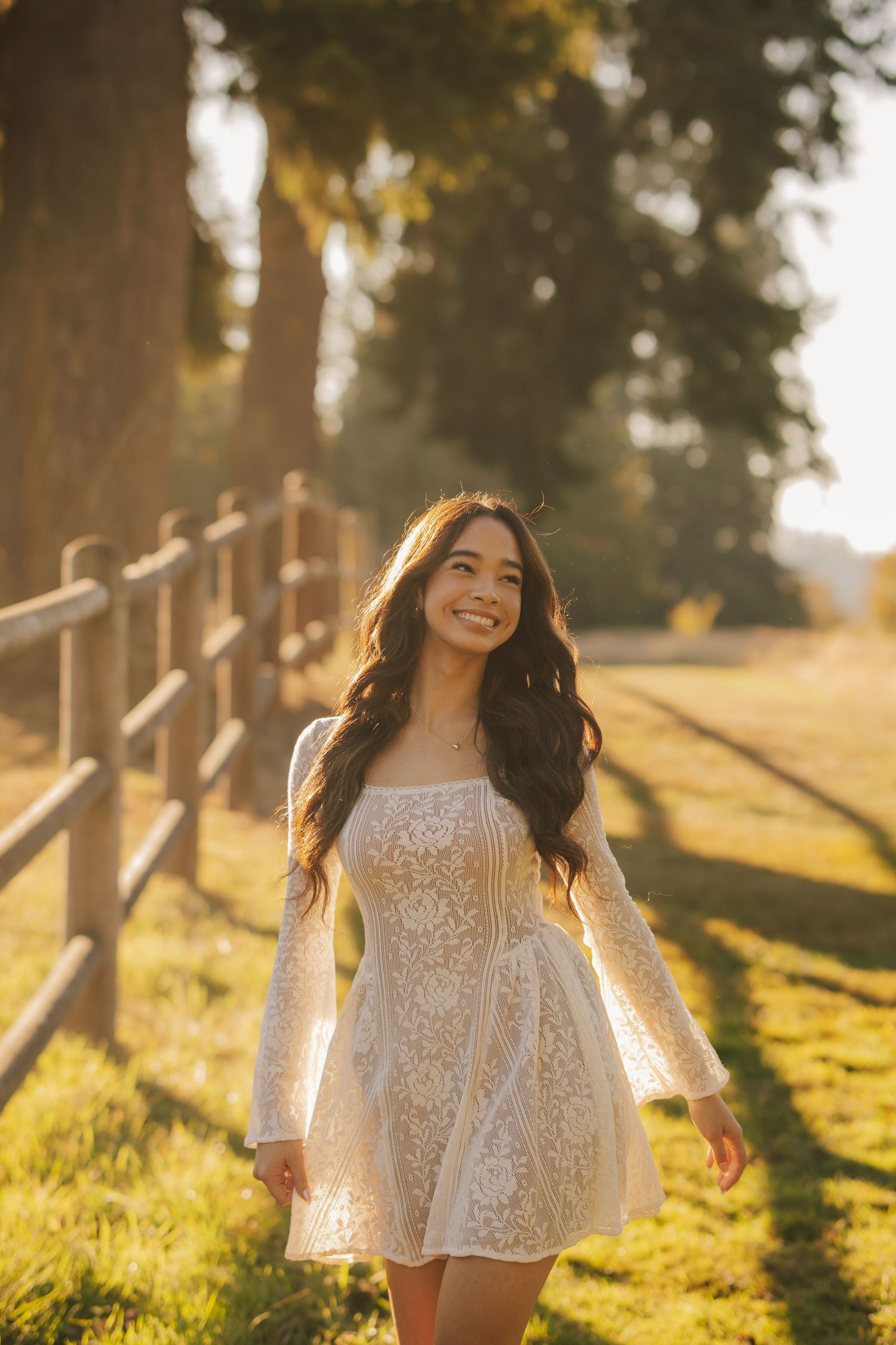 Smiling woman with long wavy dark hair in a white lace dress walks along a wooden fence on a grassy field in warm golden sunset light.