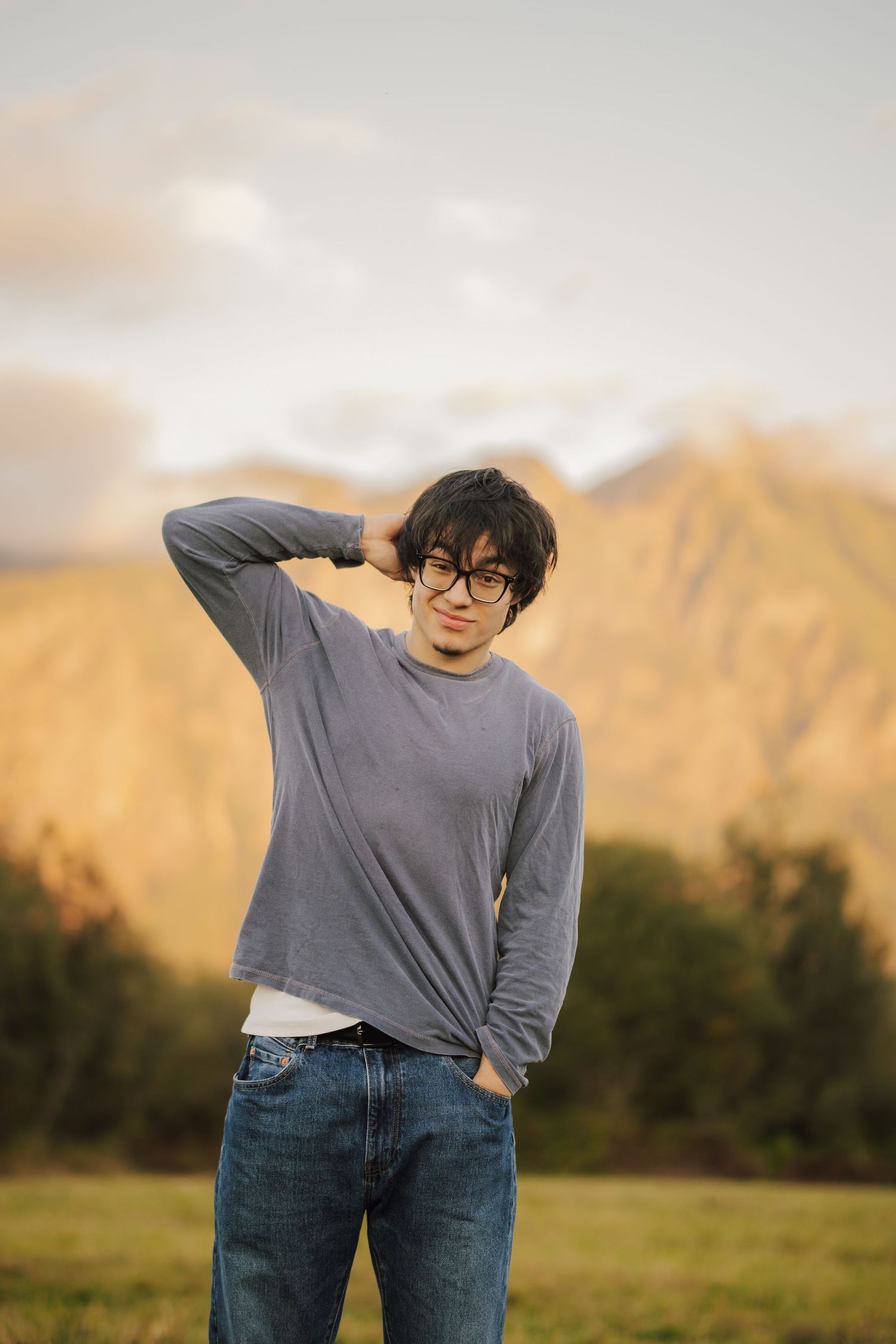 Young man with glasses and a gray long-sleeve shirt stands in a grassy field, hand behind his head, with golden mountains behind him.