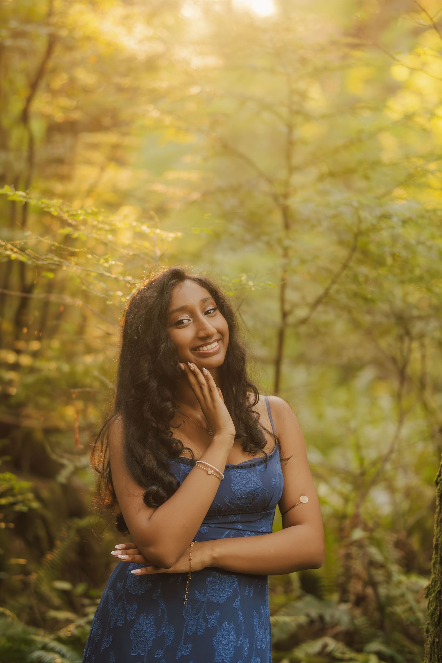 Smiling young woman with long dark wavy hair in a blue floral dress poses with her hand near her cheek in a sunlit, green forest.