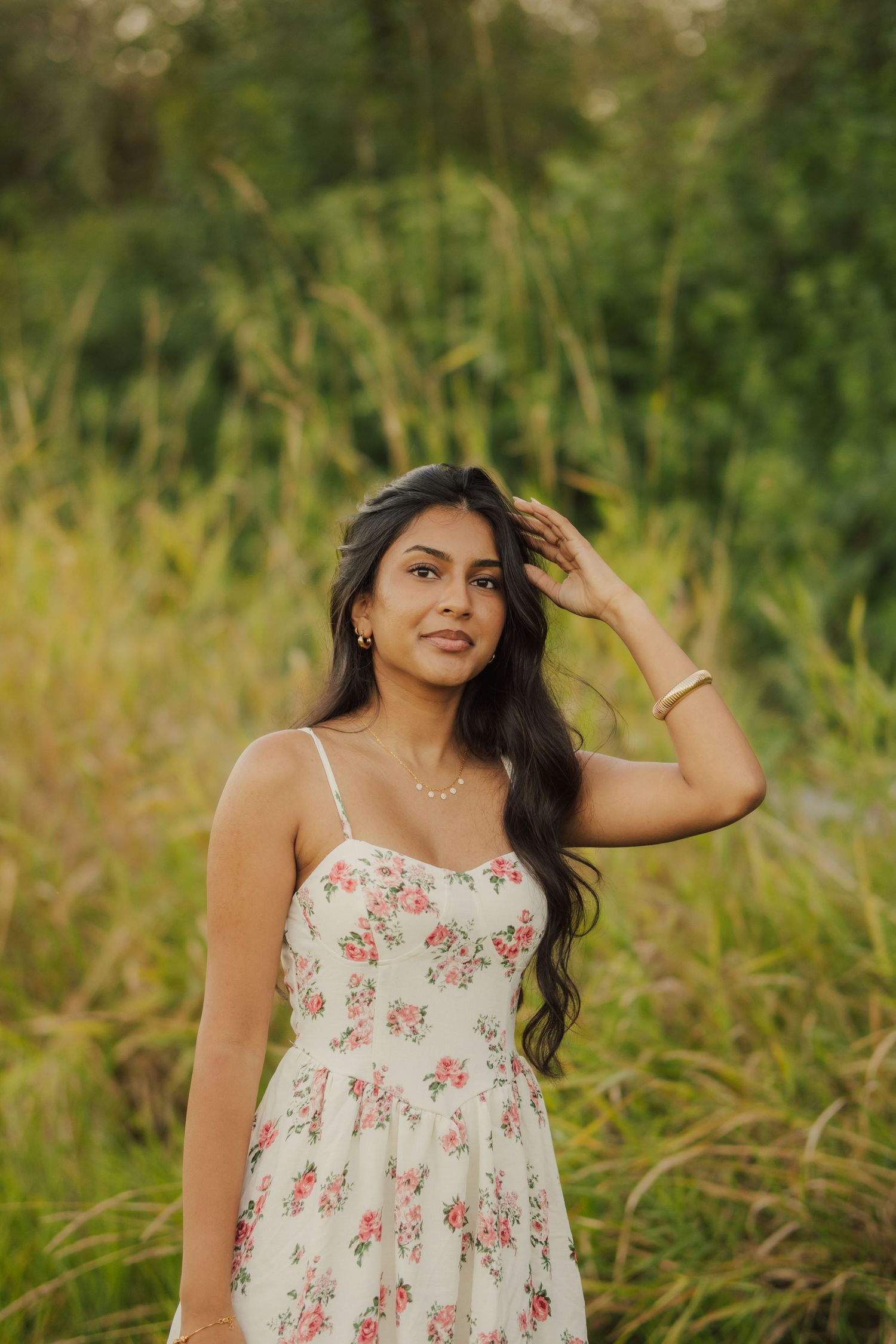 Woman with long dark wavy hair in a white floral sundress poses with her hand in her hair amid tall green grass at golden hour.