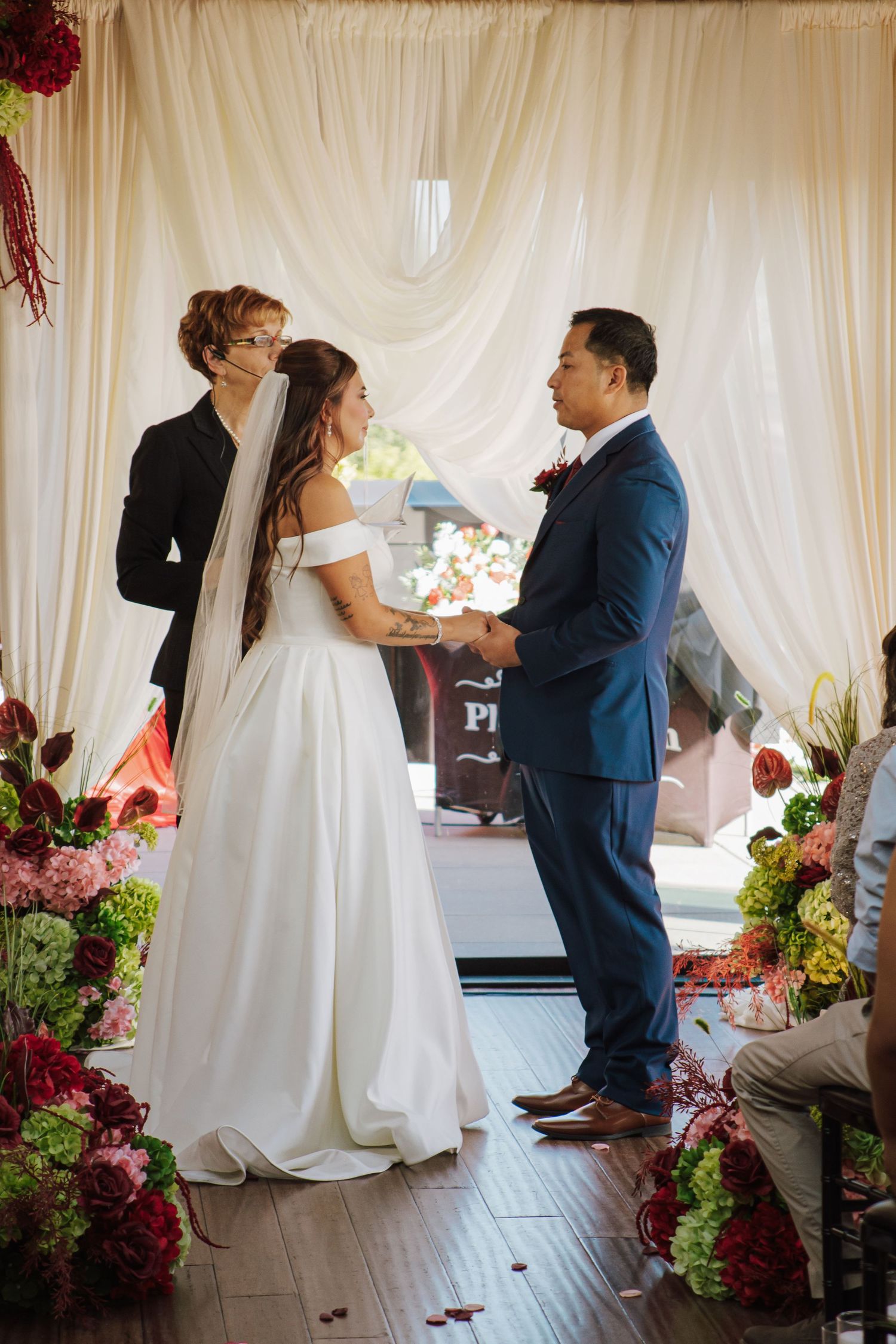 Bride and groom hold hands at altar with white draped backdrop and vibrant red and green florals.