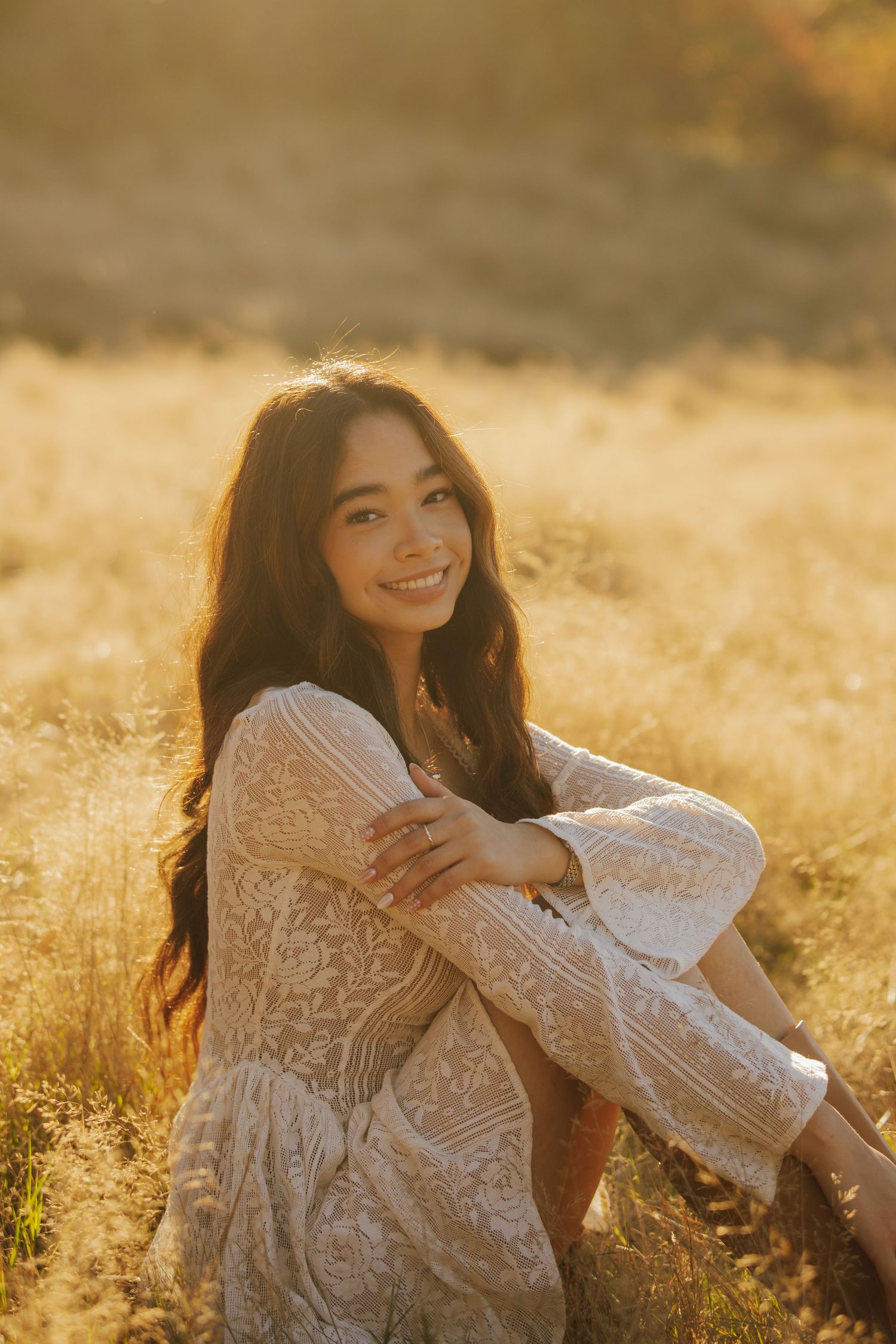 Smiling woman with long wavy dark hair in a white lace dress sits in a golden grass field, glowing in warm sunset light.