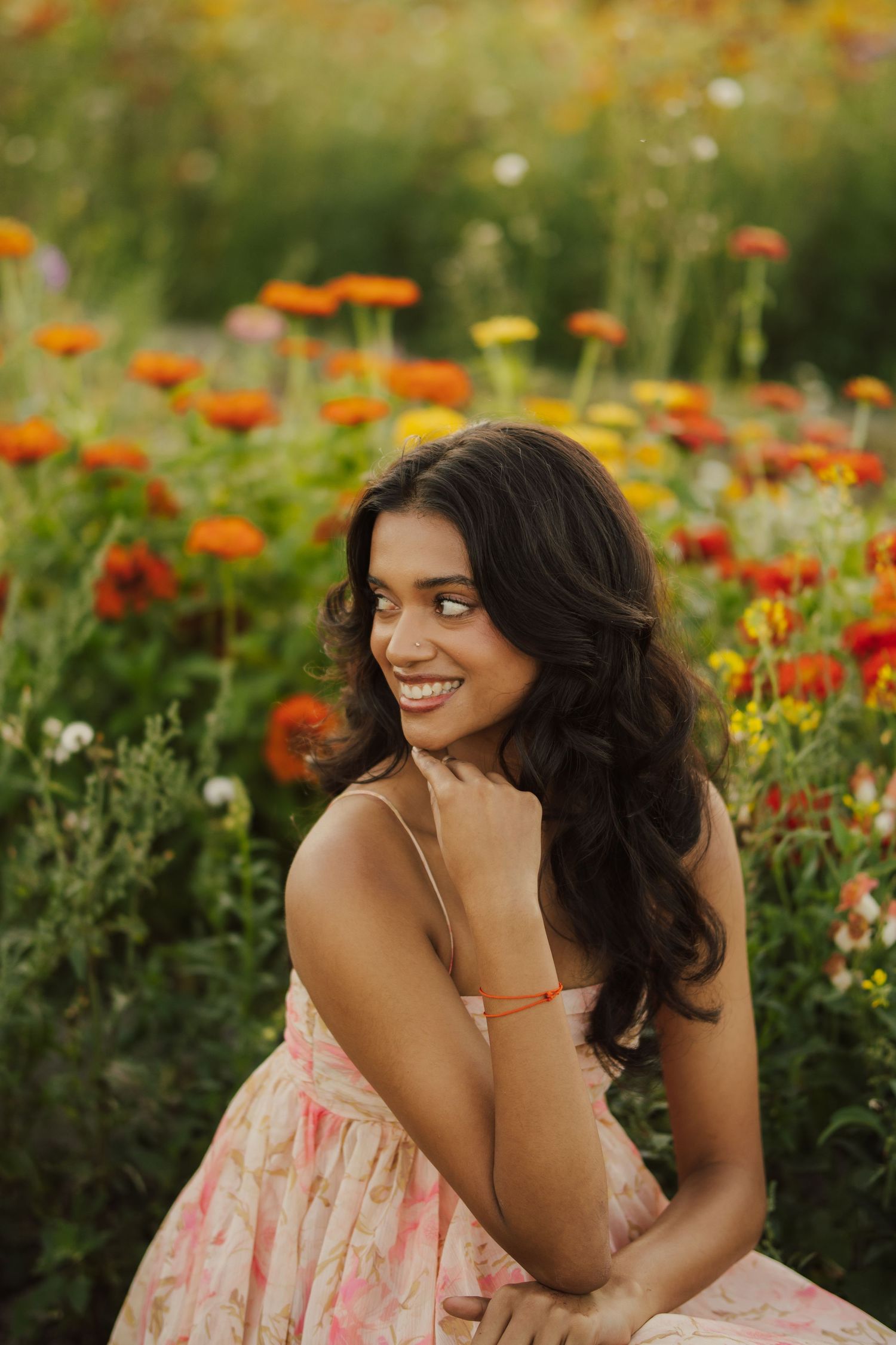 Smiling woman with long wavy dark hair in a pink floral sundress sits among bright orange and red zinnias in a sunlit flower field.