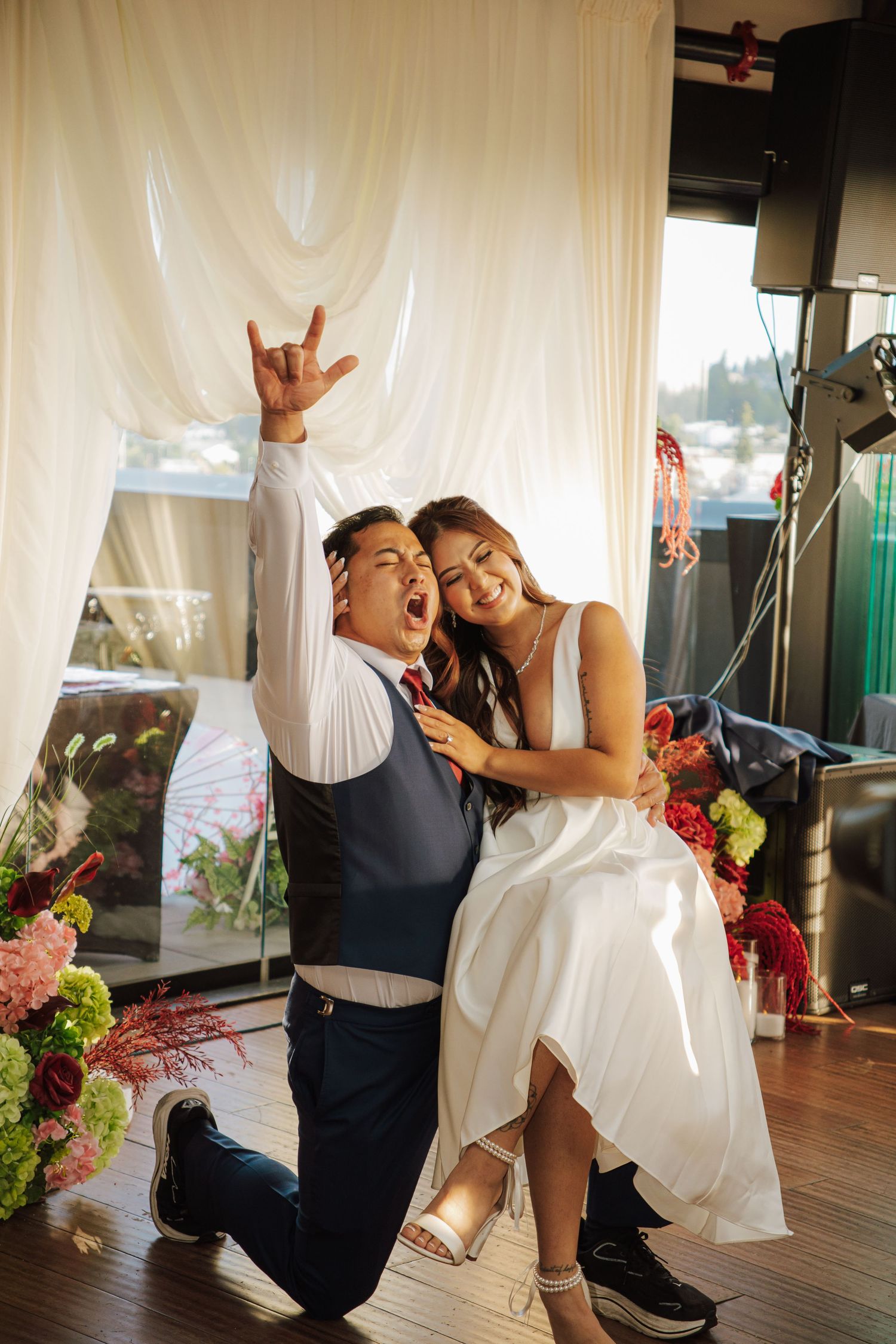 Excited groom kneels while bride sits on his knee laughing at wedding reception celebration.