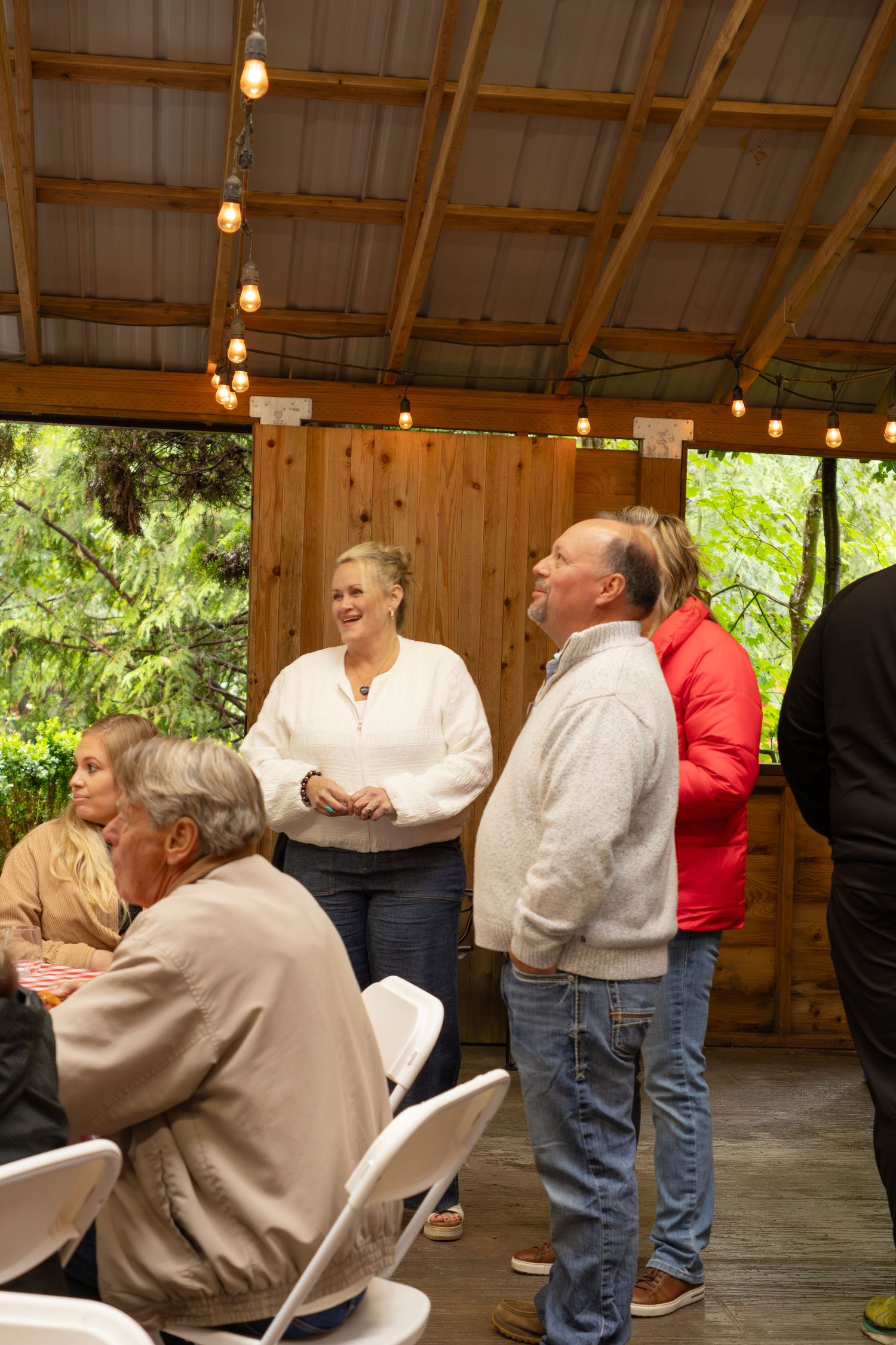 People socializing at an outdoor covered pavilion party with string lights and wooden walls.
