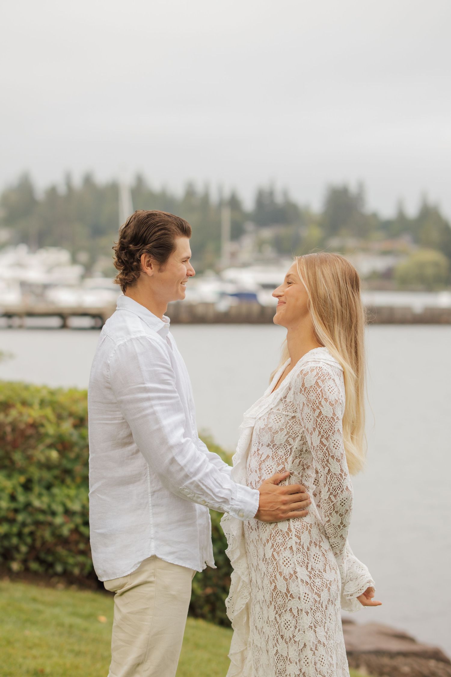 Couple in white outfits holding each other and smiling near a marina with boats in background.