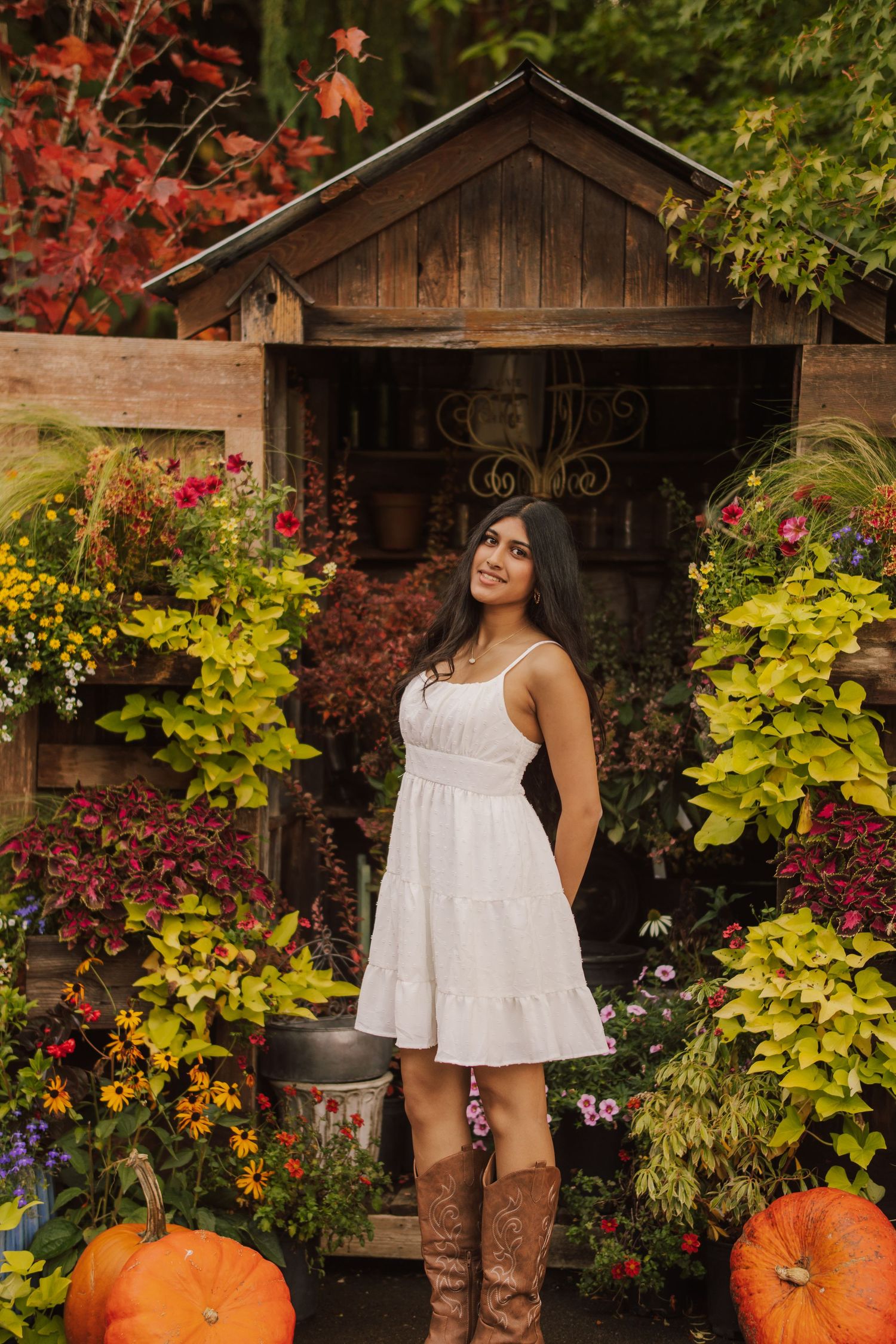 Woman in a white sundress and brown cowboy boots poses by a rustic wooden shed surrounded by colorful autumn flowers and orange pumpkins.
