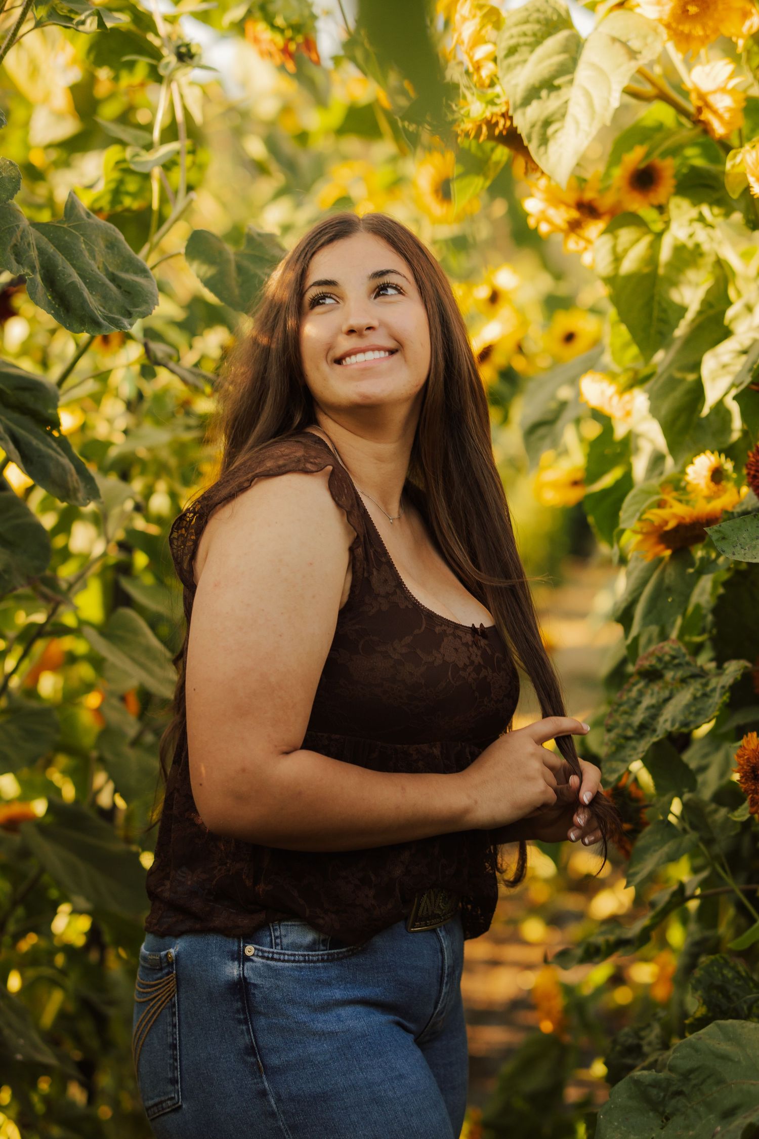 Smiling woman with long brown hair in a dark lace tank top and jeans stands among sunflowers in warm golden sunlight.