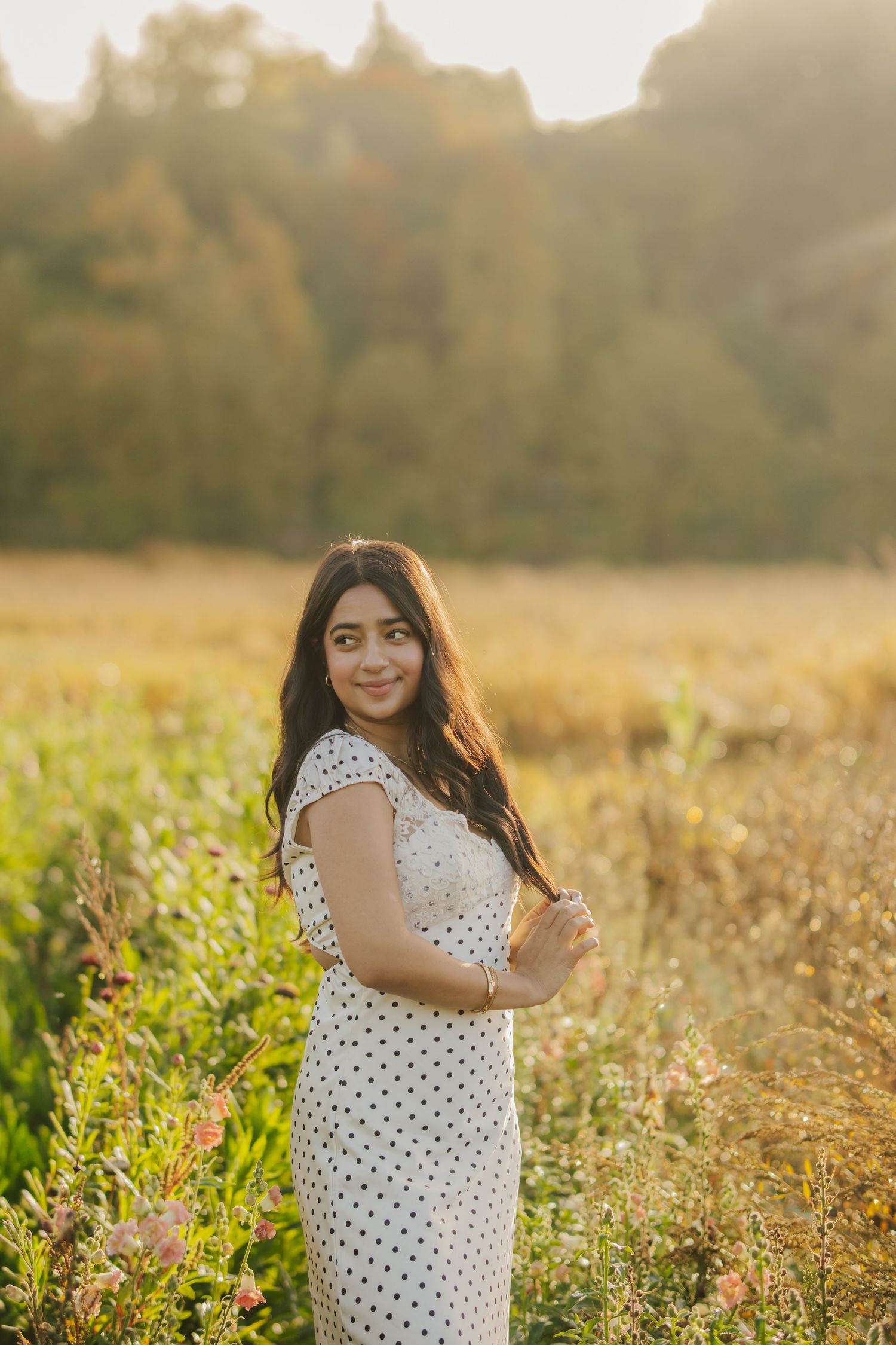 Smiling woman with long dark hair in a white polka dot dress poses in a golden meadow with wildflowers and trees in warm sunset light.
