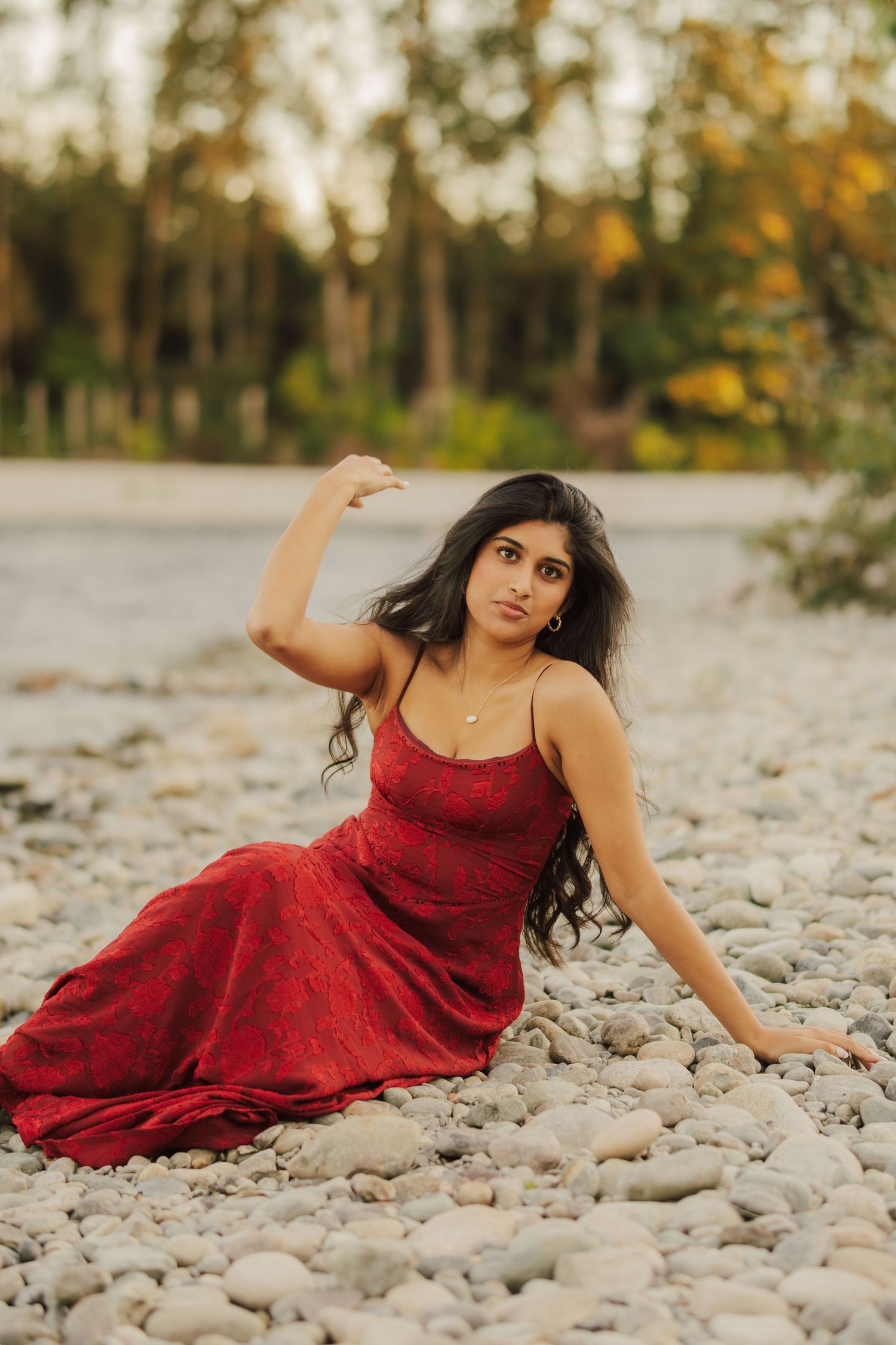 Woman in a flowing red dress sits on a smooth pebble riverbank with one arm raised, framed by autumn trees in the soft background.