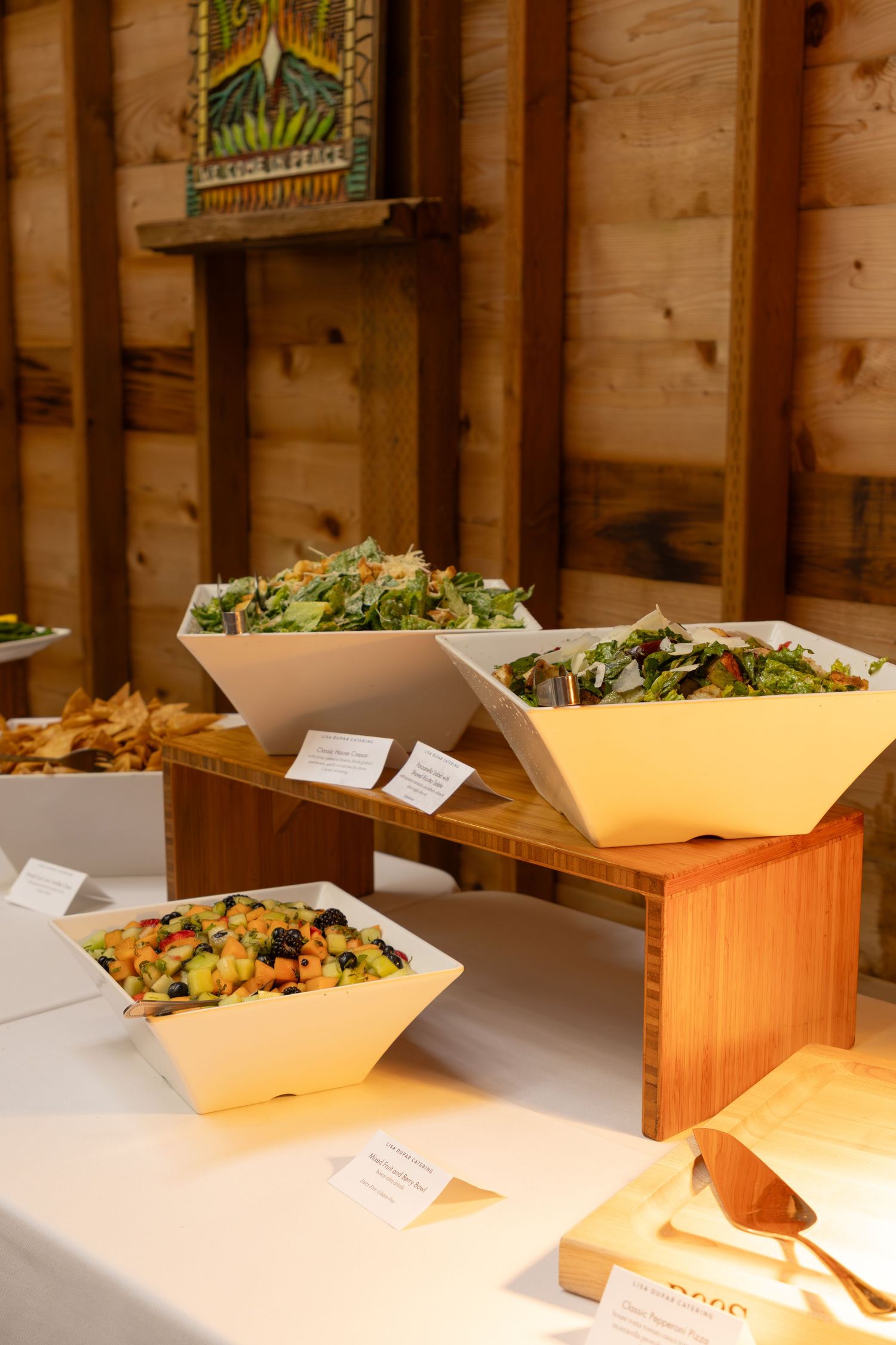 Buffet table with salad bowls on wooden risers at an indoor event with string lights.
