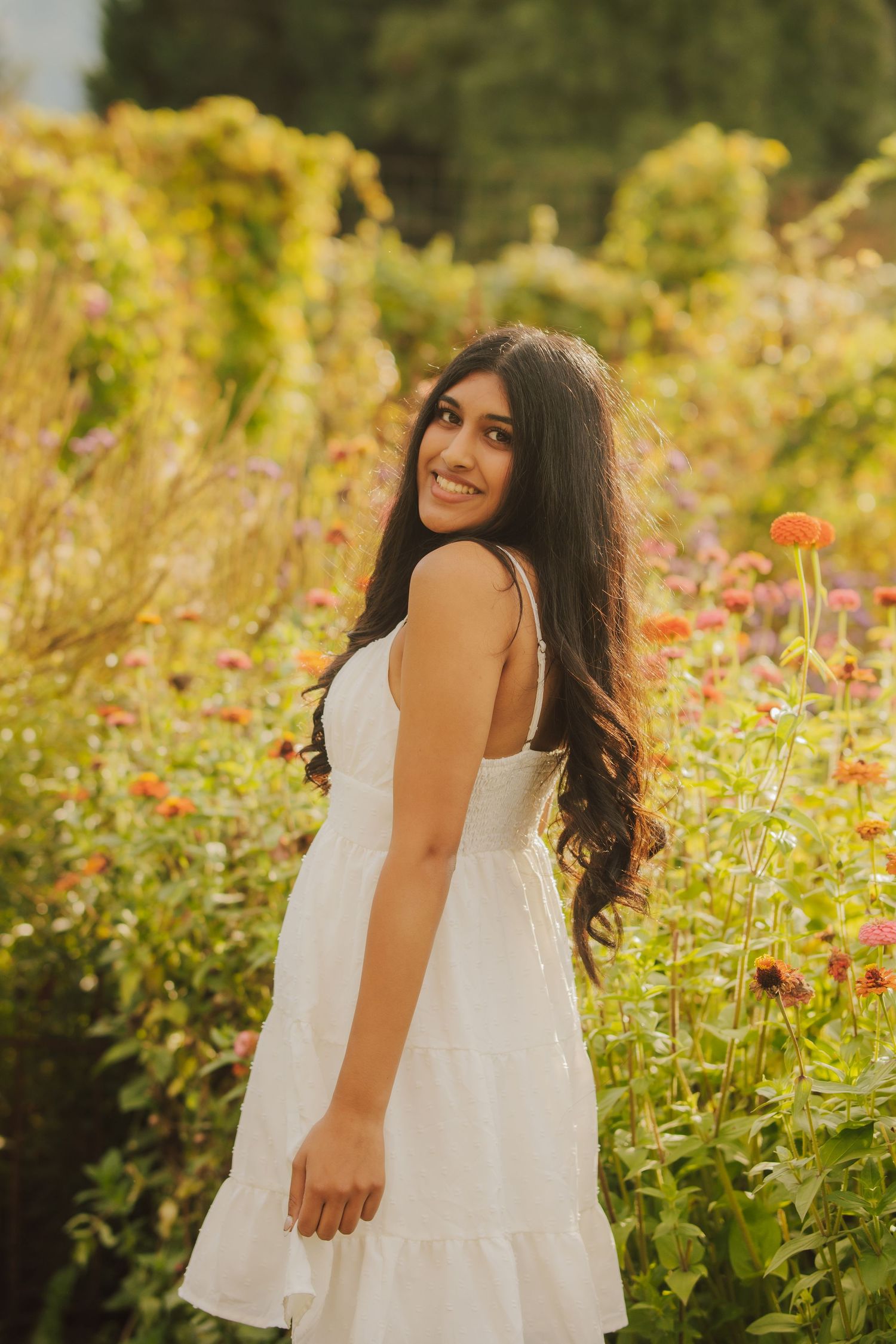 Smiling woman with long dark wavy hair in a white tiered sundress stands among blooming orange and pink zinnias in a sunlit garden.