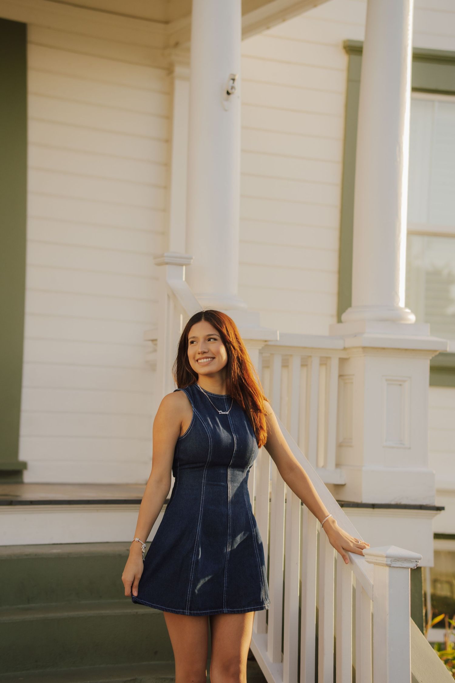 Smiling young woman in a sleeveless denim dress stands on porch steps beside a white railing and columns of a clean white house.