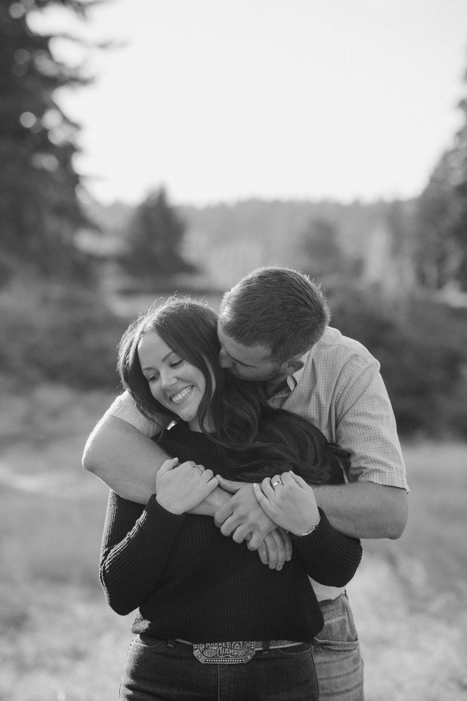 Black and white engagement photo of a smiling woman hugged from behind by a man kissing her cheek in a field with trees.