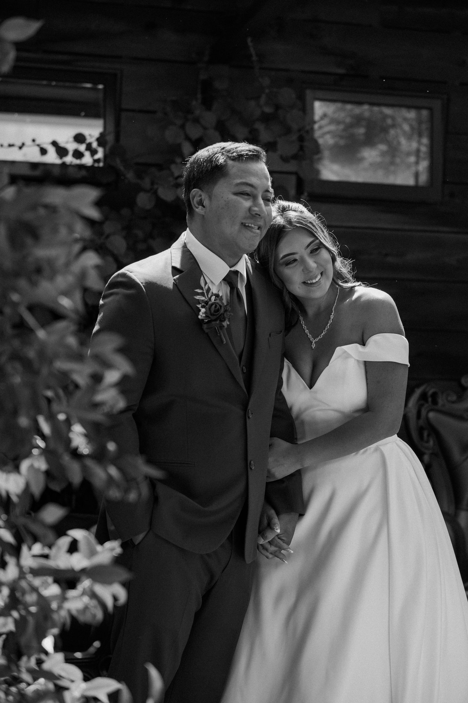 Black and white portrait of bride and groom embracing in front of a rustic cabin.