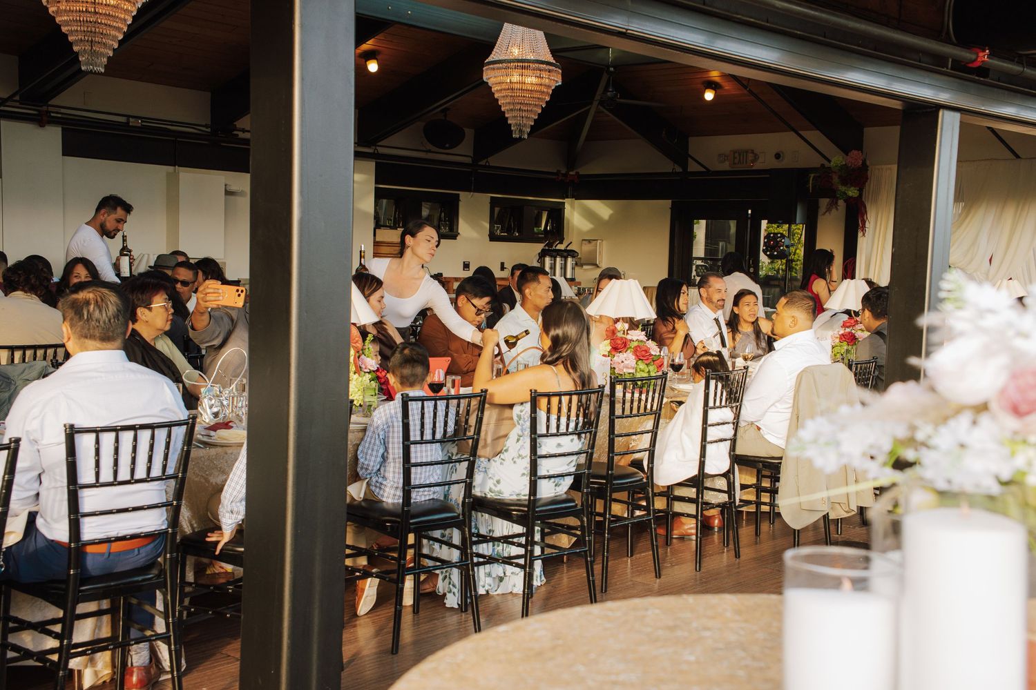 Wedding reception guests seated at decorated tables inside an elegant venue with chandelier lighting.