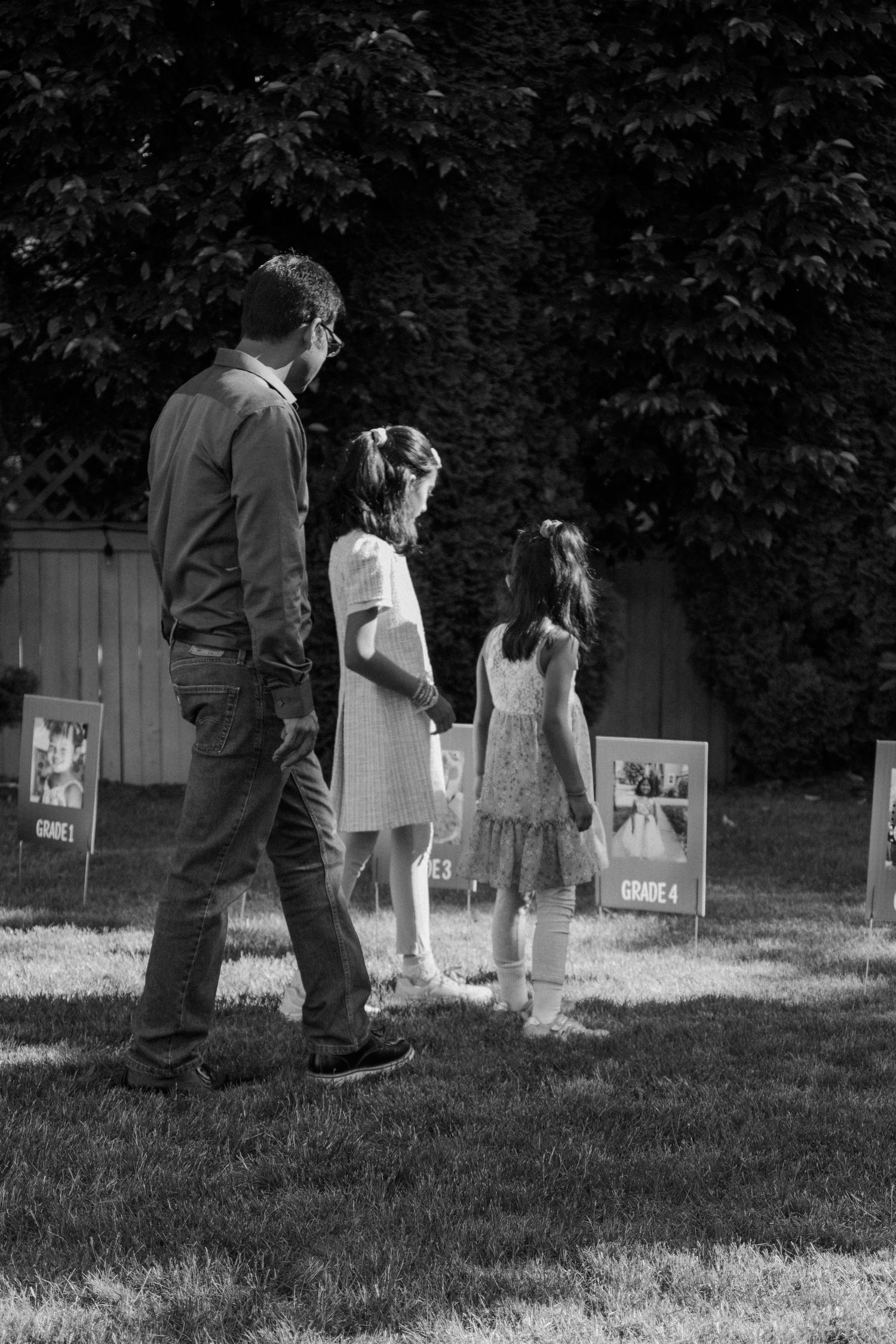 Black and white photo of father walking with two young girls viewing grade school photo displays.