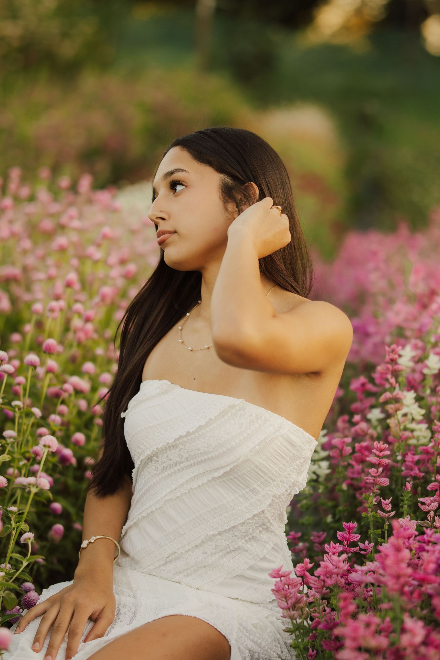 Young woman with long dark hair in a white off-shoulder dress sits among blooming pink flowers, hand raised to her hair at golden hour.