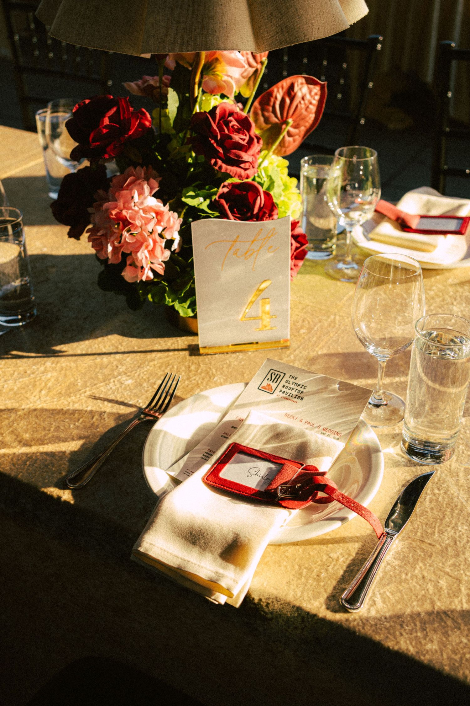 Elegant wedding reception place setting with red floral centerpiece, glassware, and menu card in sunlight.