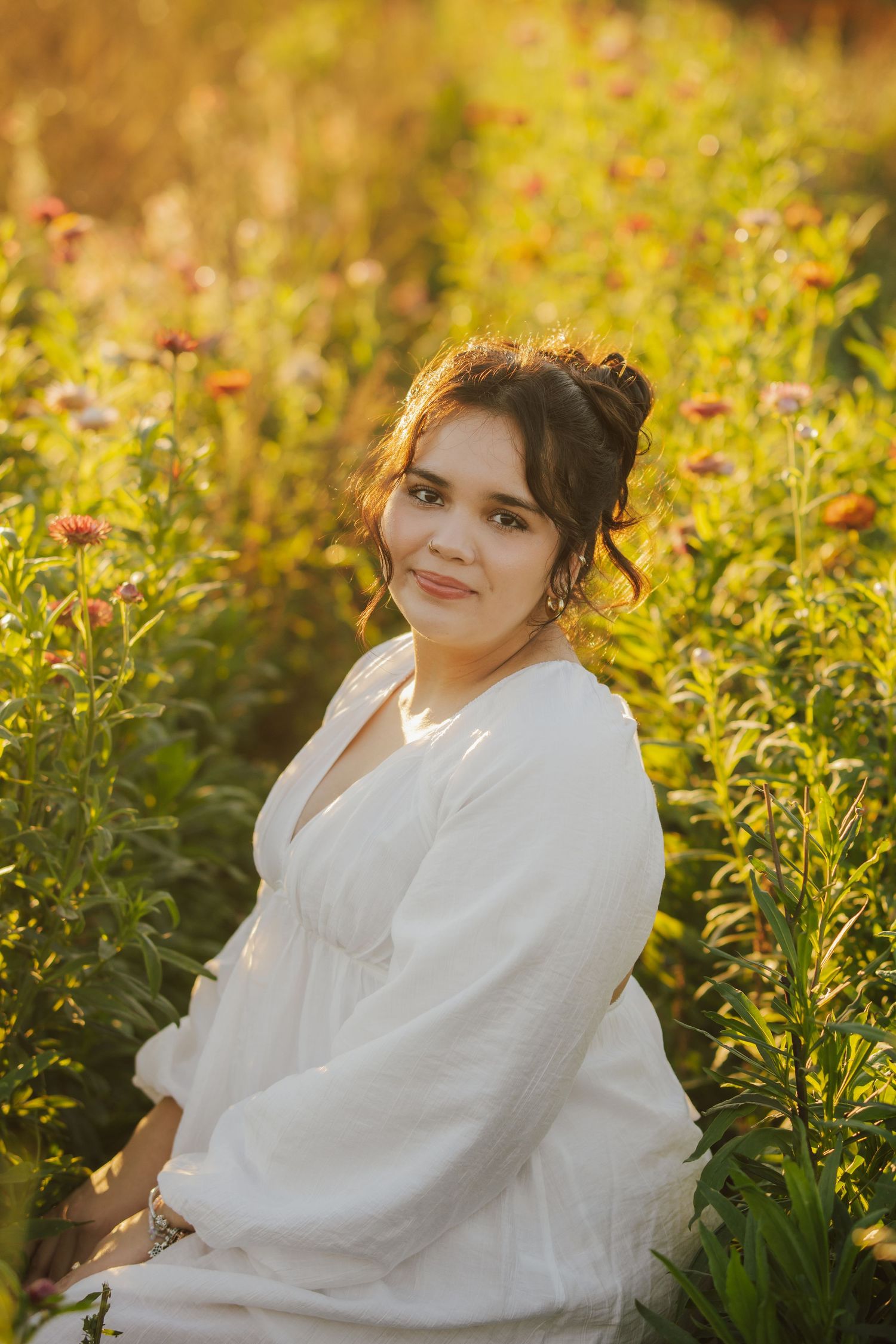 Smiling woman in a white flowy dress sits among blooming pink and orange wildflowers in a field, glowing in warm golden-hour sunlight.