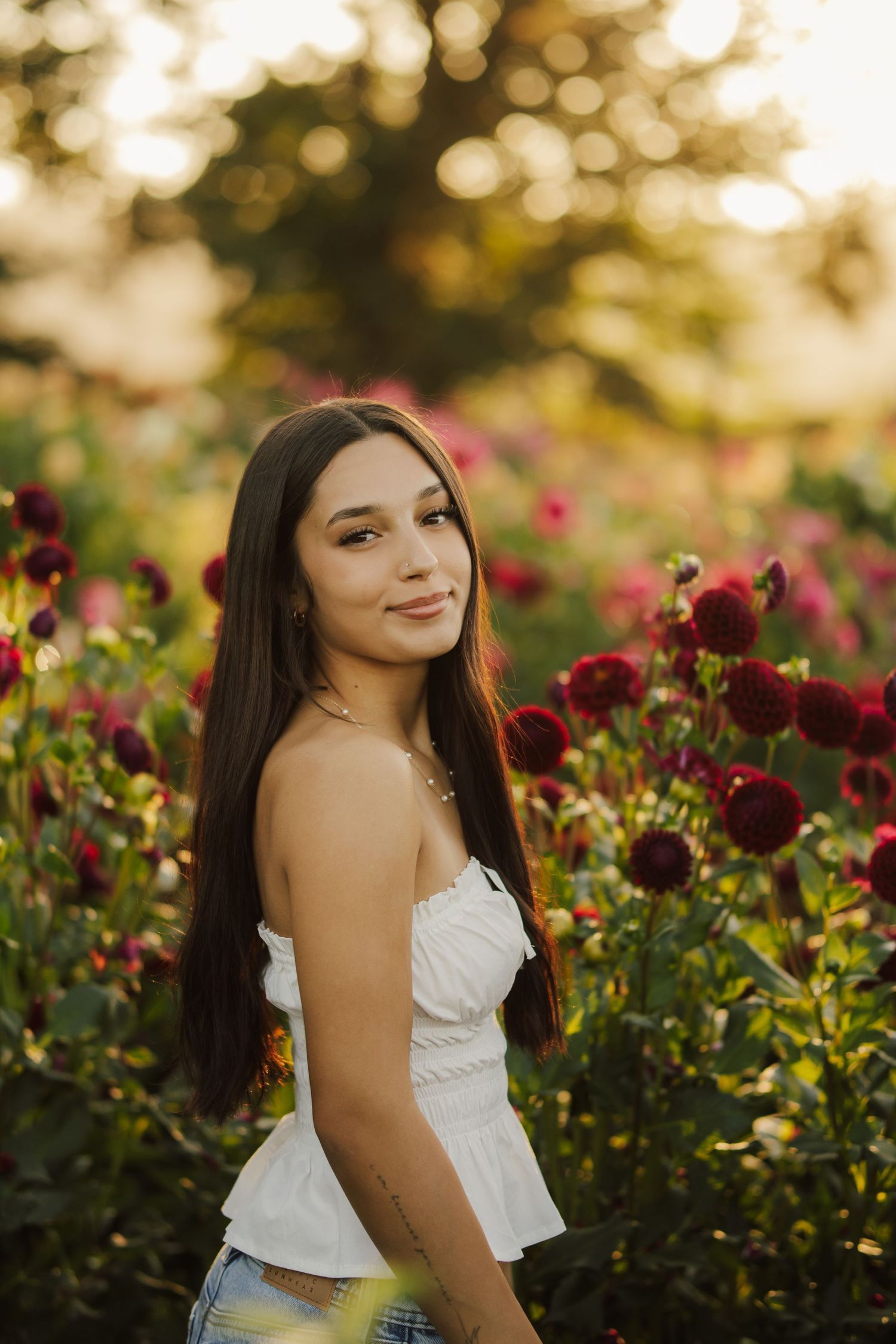 Young woman with long dark hair in a white strapless top poses among blooming red dahlias in a flower field at golden hour.