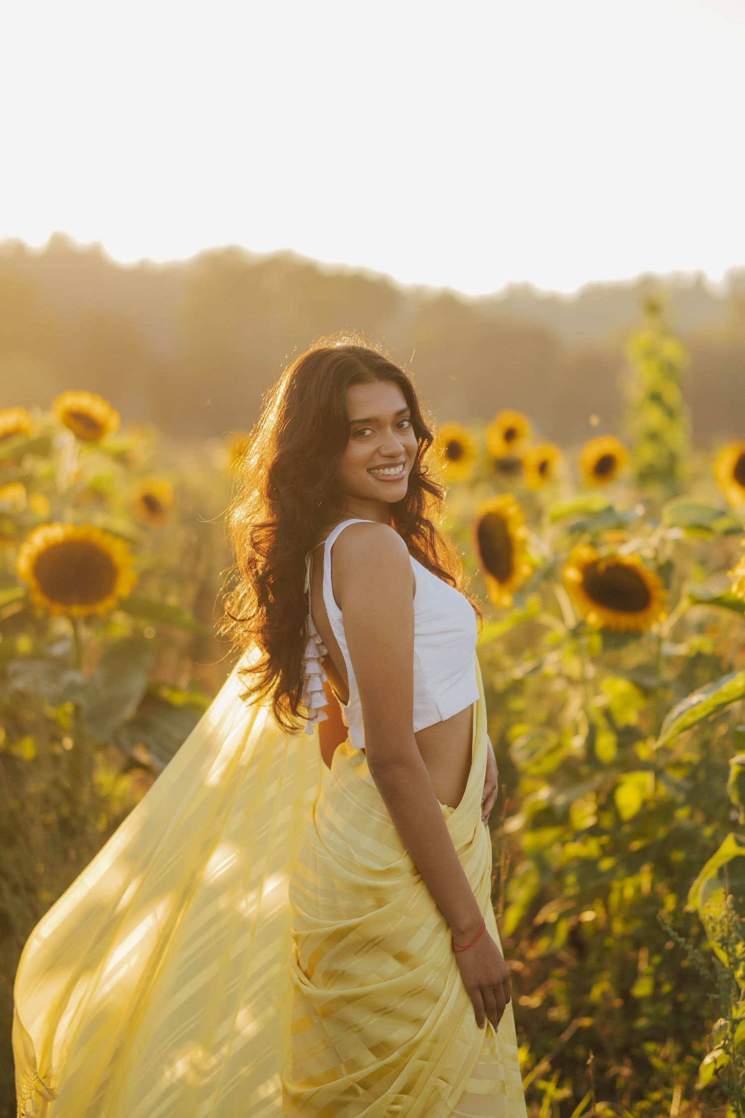 Smiling woman with long wavy hair in a flowing yellow saree stands in a sunflower field at golden hour, glancing back over her shoulder.