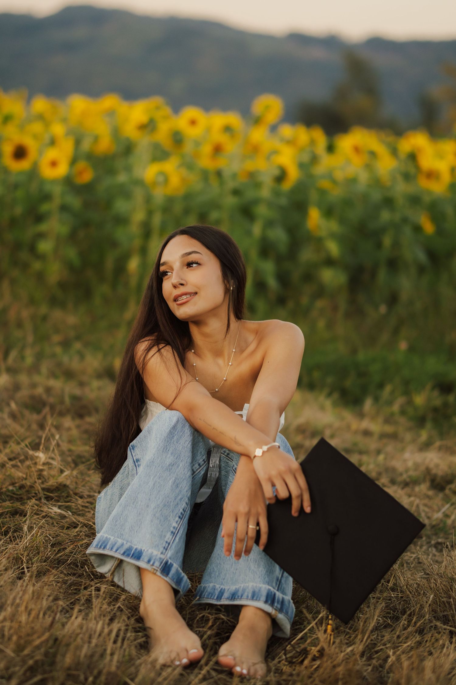 Young woman with long dark hair sits barefoot in a grassy field of sunflowers, holding a black graduation cap at golden hour.
