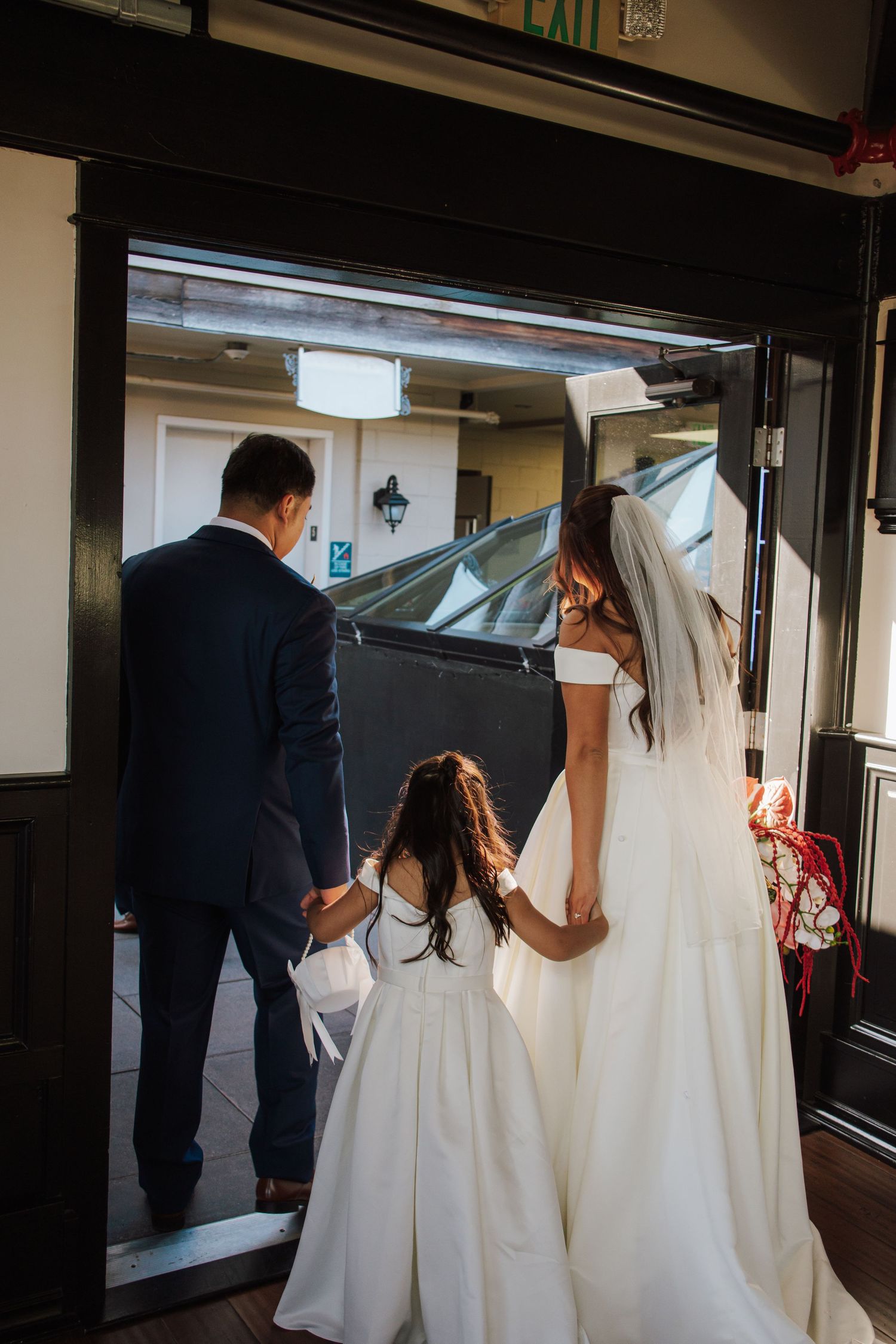 Bride in white gown and veil walks with groom and flower girl through venue doorway at wedding.