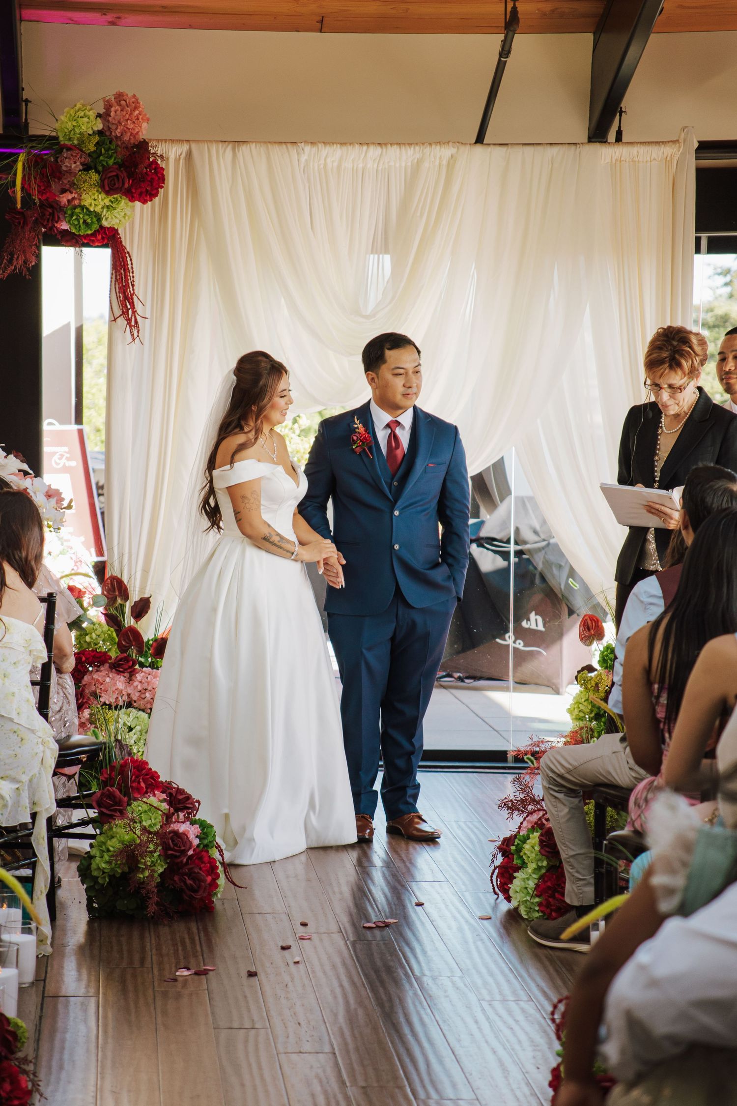 Bride and groom walk down aisle together in decorated indoor wedding venue with white drape backdrop.