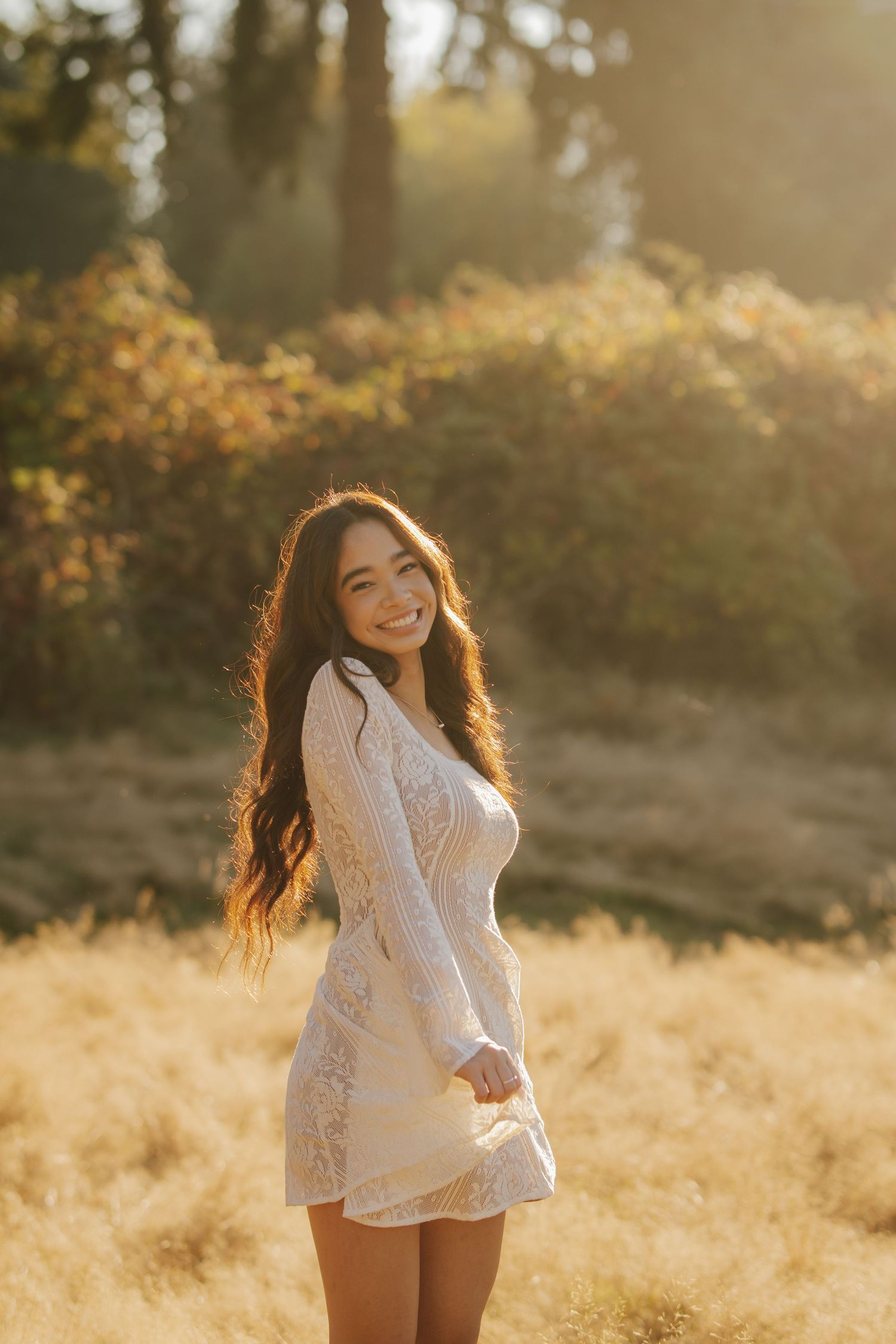 Smiling woman with long wavy dark hair in a white lace dress stands in a golden grass field at sunset surrounded by warm glowing trees.