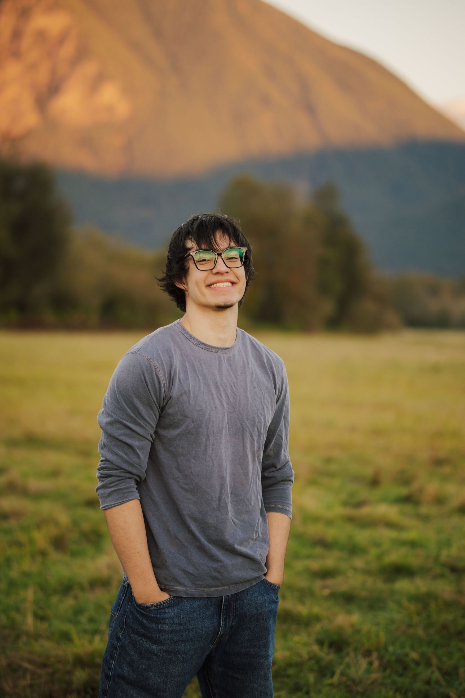 Smiling young man in green glasses and a gray sweater stands with hands in pockets in a grassy field before a sunlit mountain.