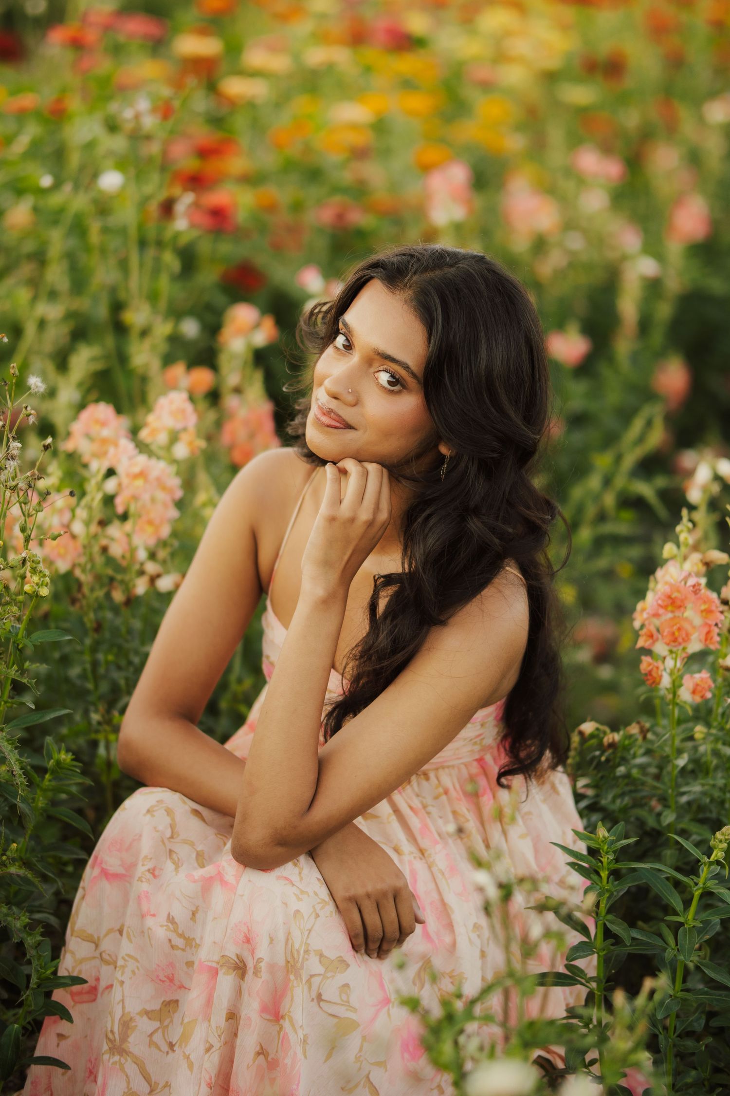 Woman with long dark wavy hair in a pink floral sundress poses with her chin on her hand amid a colorful blooming flower field.