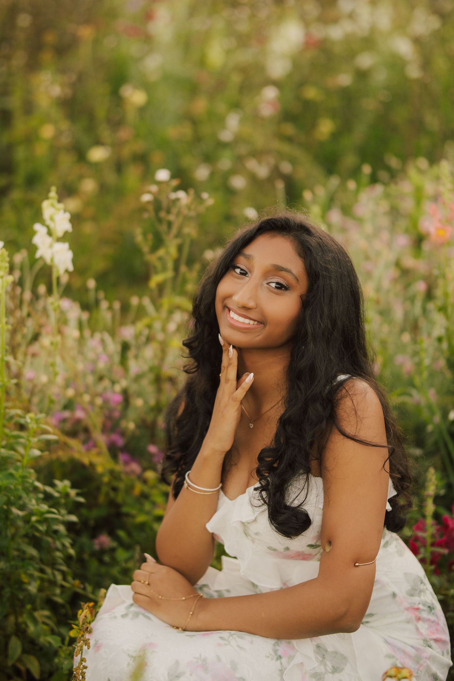 Smiling young woman with long dark wavy hair in a floral sundress poses with her hand on her chin in a colorful wildflower garden.