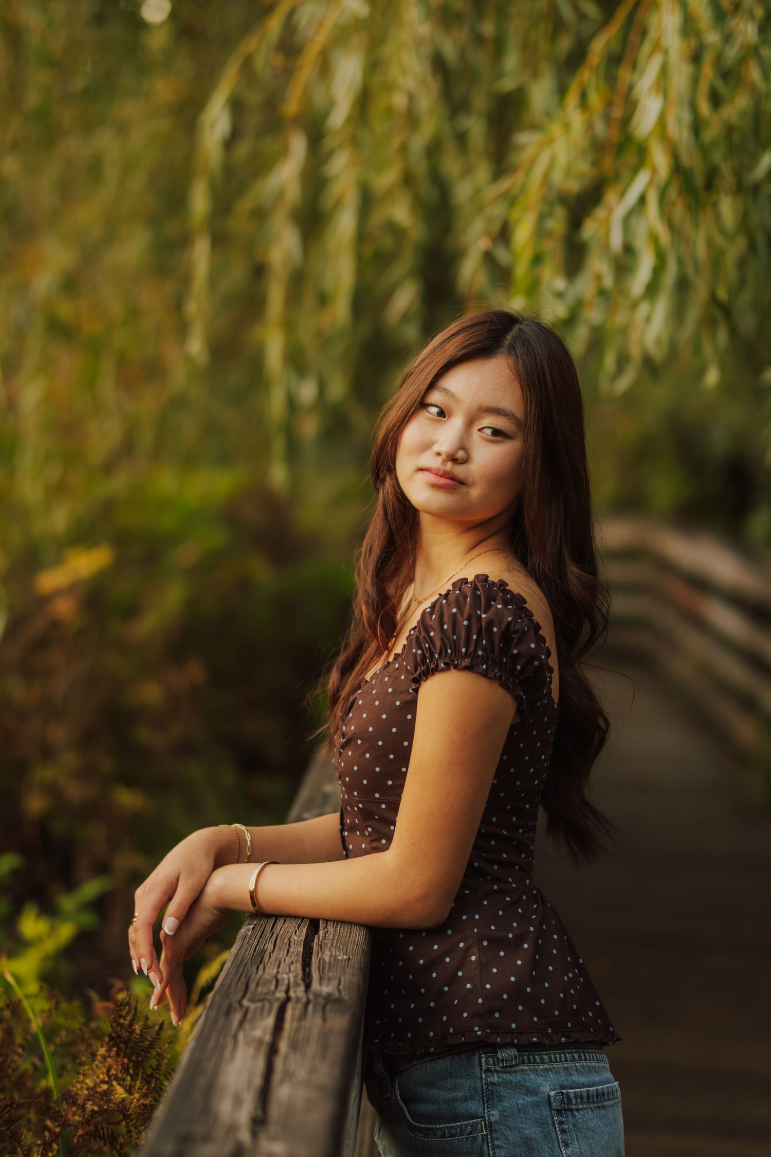 Young woman with long brown hair in a polka dot top leans on a wooden railing beneath weeping willow branches in warm golden light.