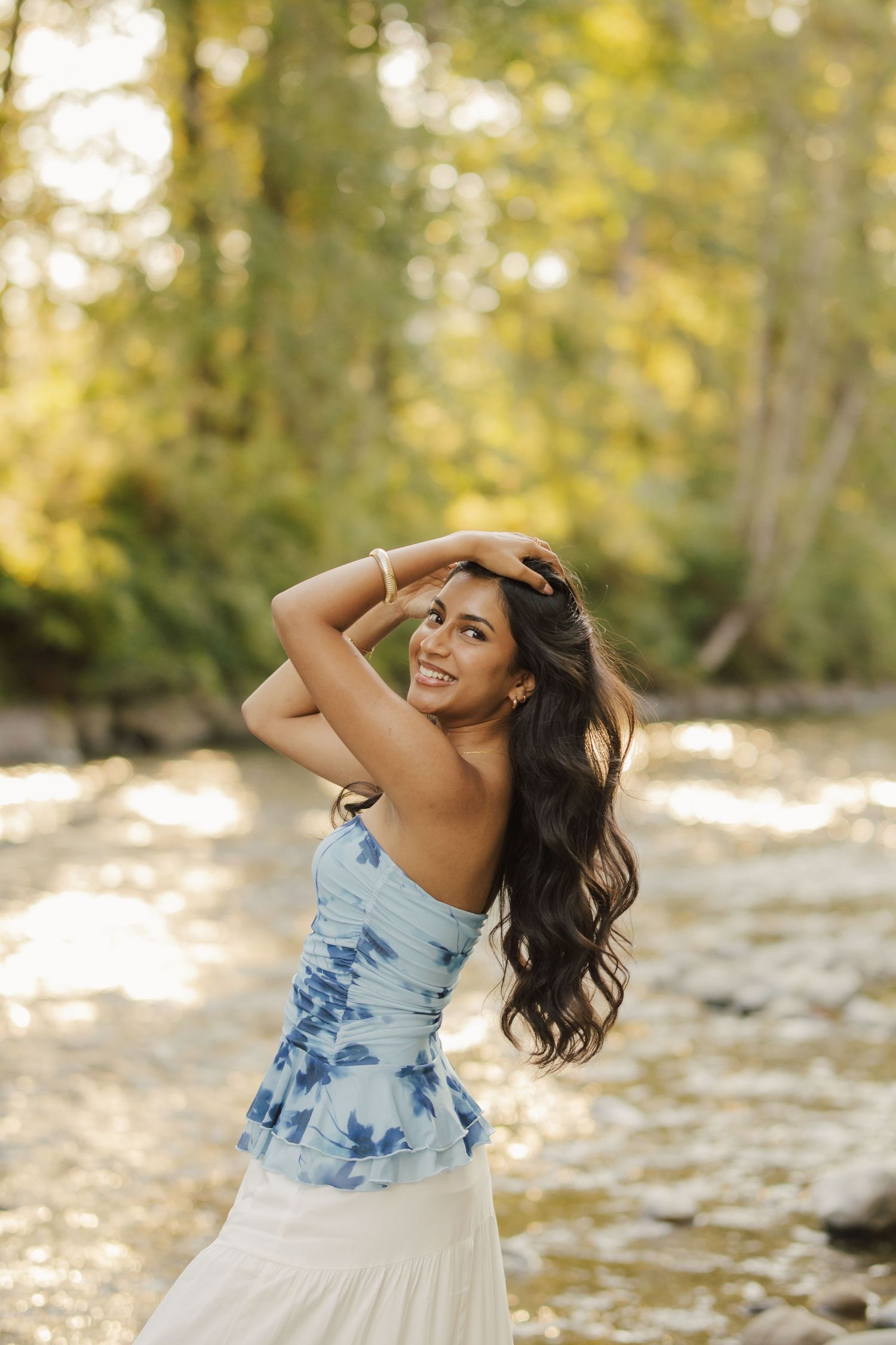 Smiling woman with long dark wavy hair poses with arms raised near a sunlit stream, wearing a blue floral top and white skirt.