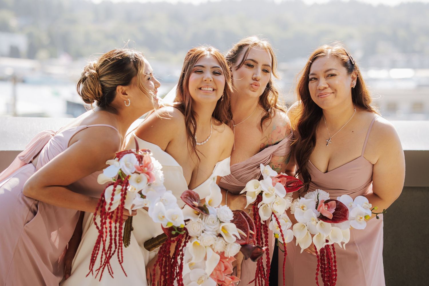 Four bridesmaids in blush pink dresses pose playfully holding white orchid and red bouquets at outdoor wedding.