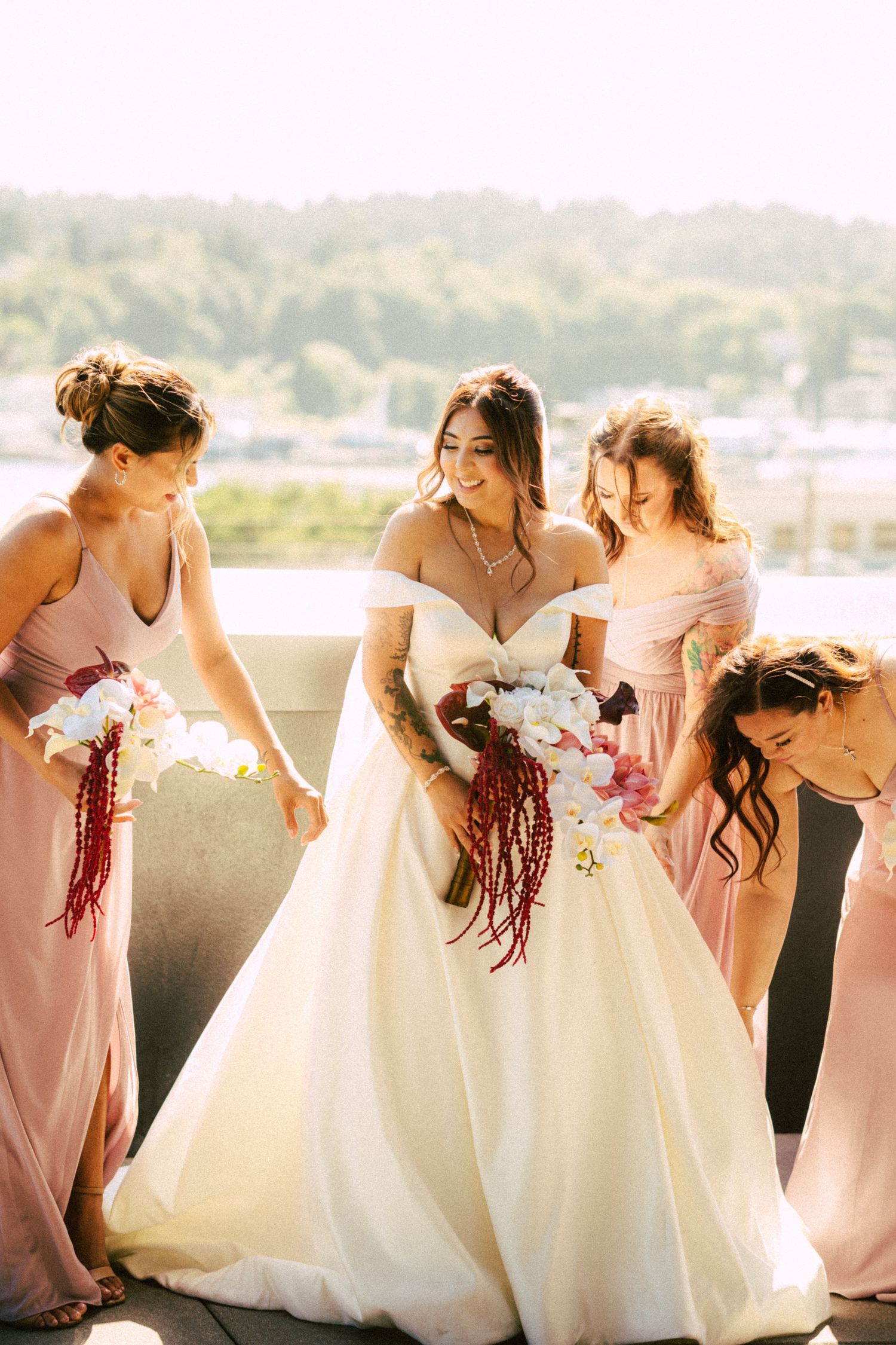 Bride in white off-shoulder gown smiles with three bridesmaids in pink dresses holding floral bouquets outdoors.