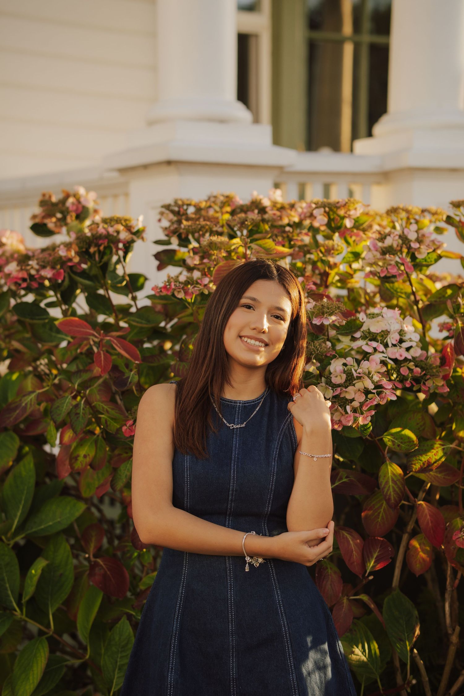 A smiling young woman in a sleeveless denim dress poses beside blooming pink hydrangeas and green leaves in warm golden sunlight.