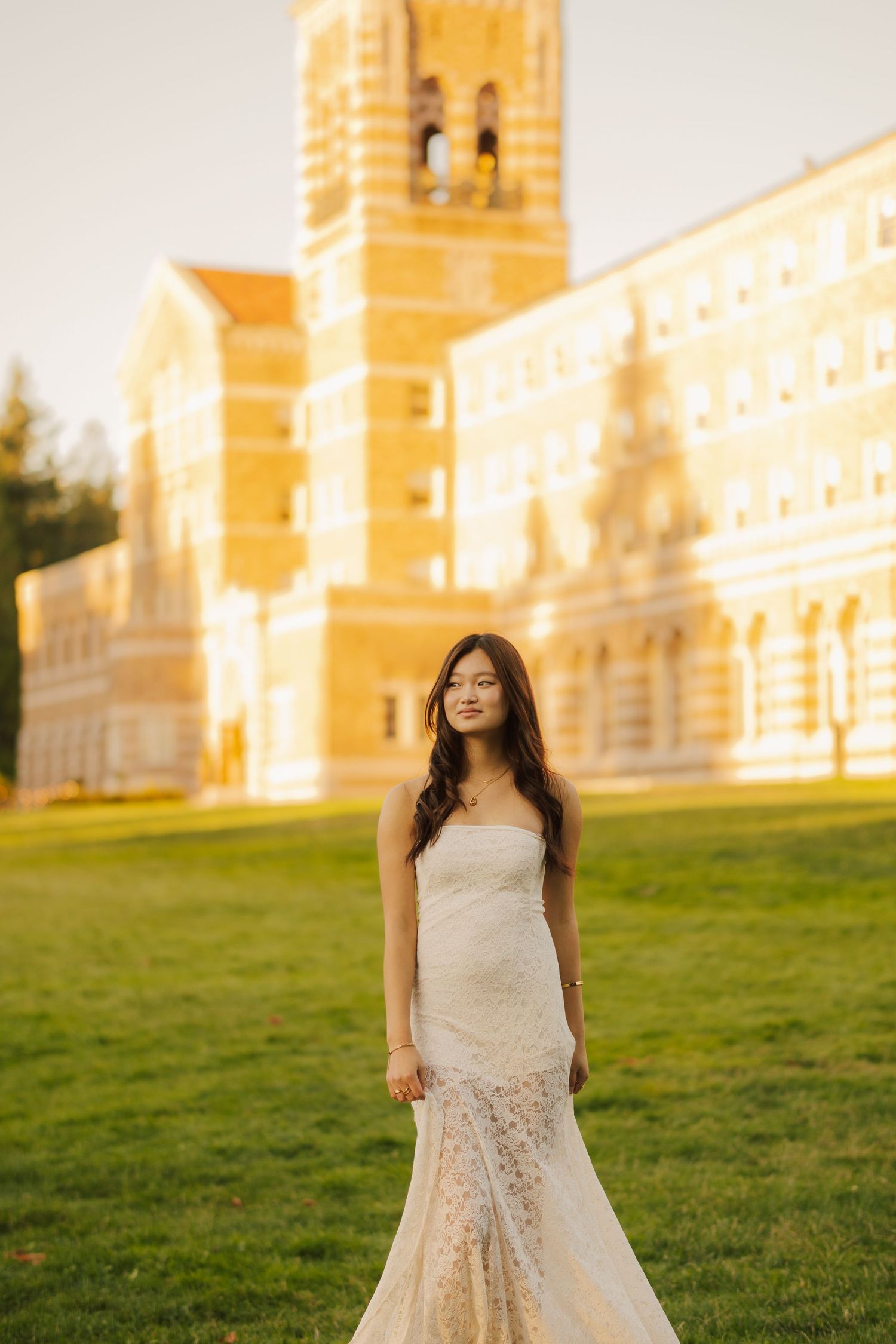 Woman in a white lace strapless gown stands on a sunlit green lawn in front of a historic tan brick building with a bell tower.