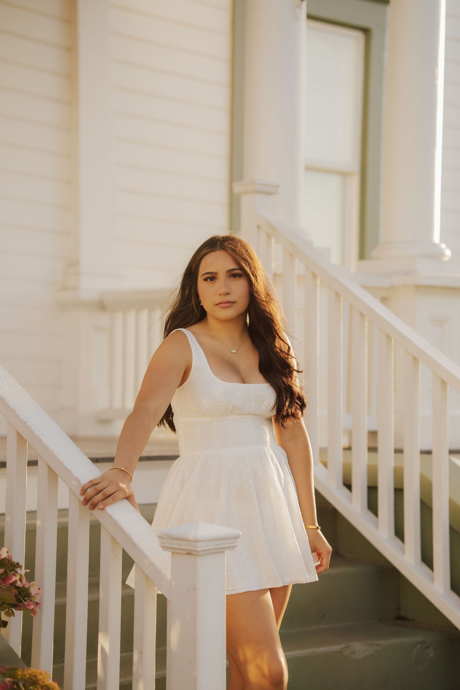 Young woman with long wavy brown hair in a white sundress poses on white porch stairs in warm golden-hour light.