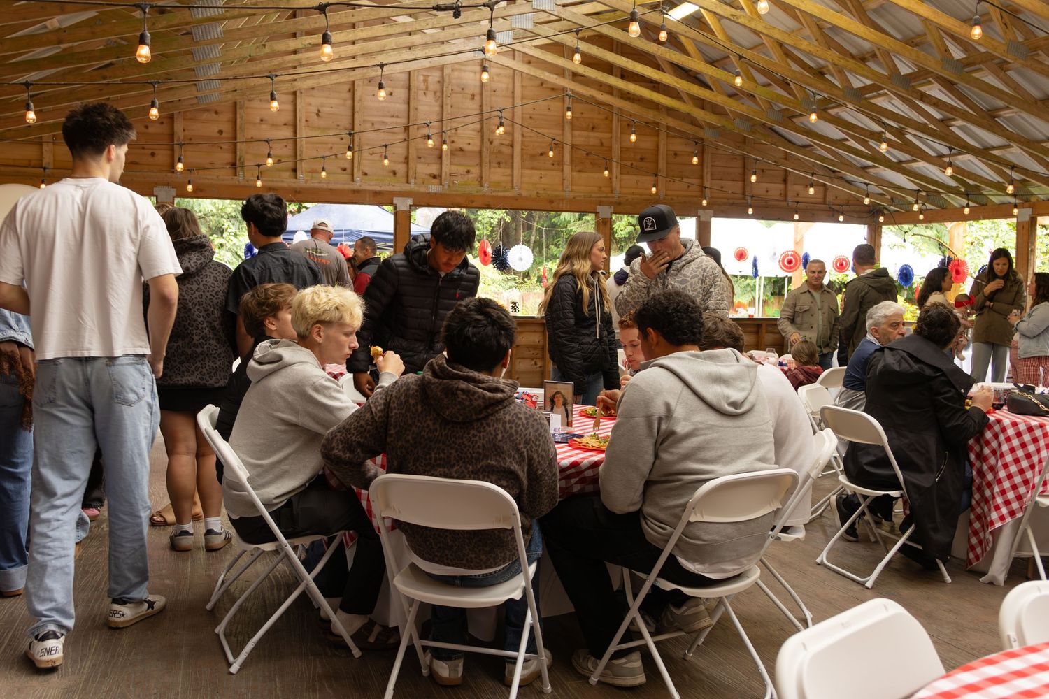 Crowd of guests dining and mingling under a wooden pavilion decorated with string lights.