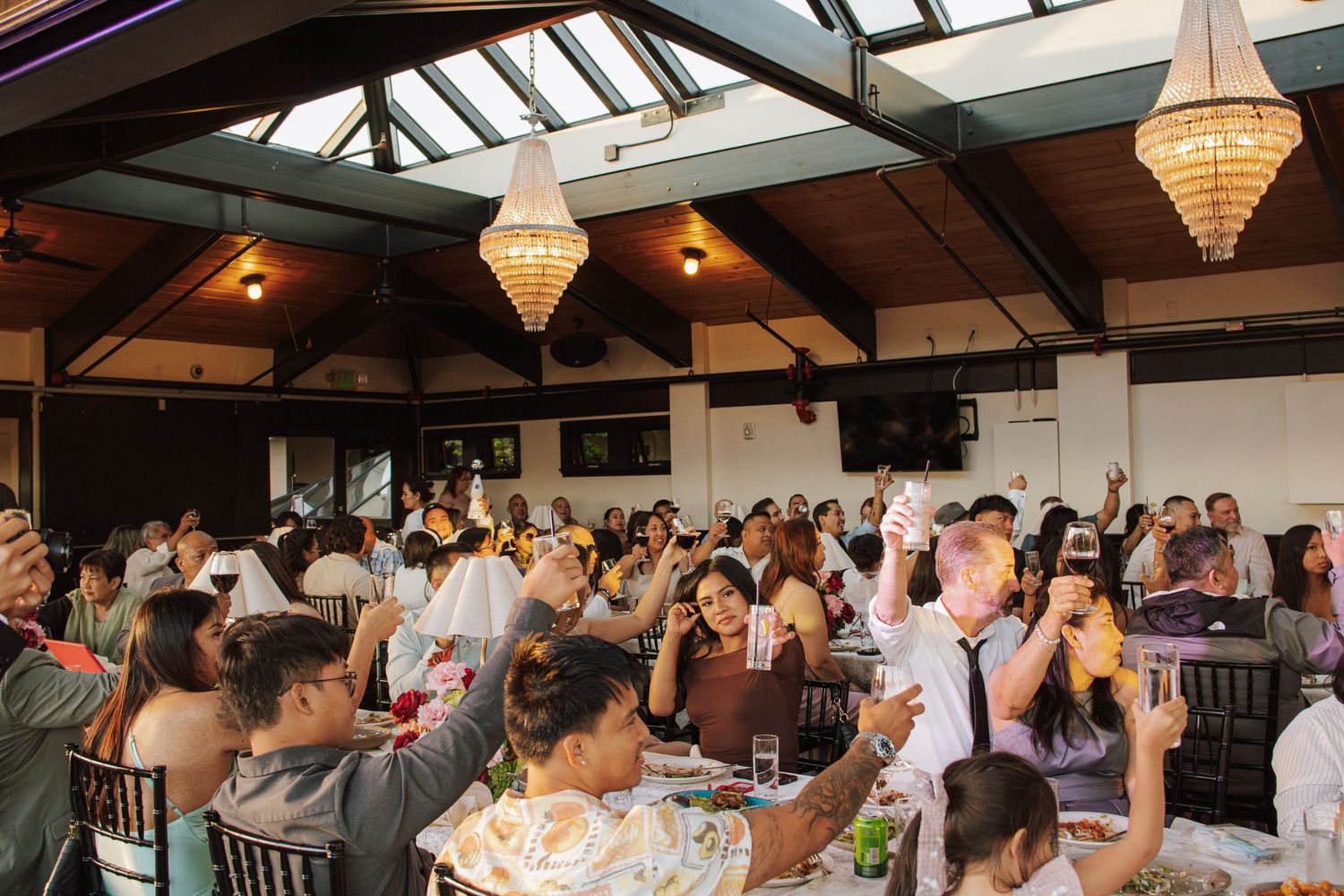 Guests raise glasses in toast at crowded wedding reception under chandelier-lit venue.