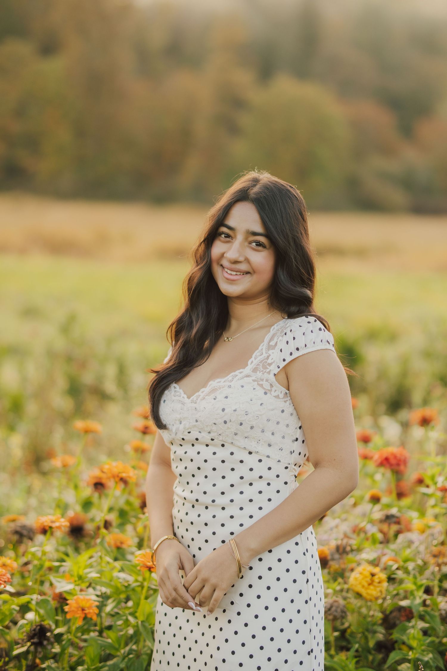 Smiling woman with long dark wavy hair wearing a white polka dot dress stands in a golden field of orange zinnia flowers at sunset.