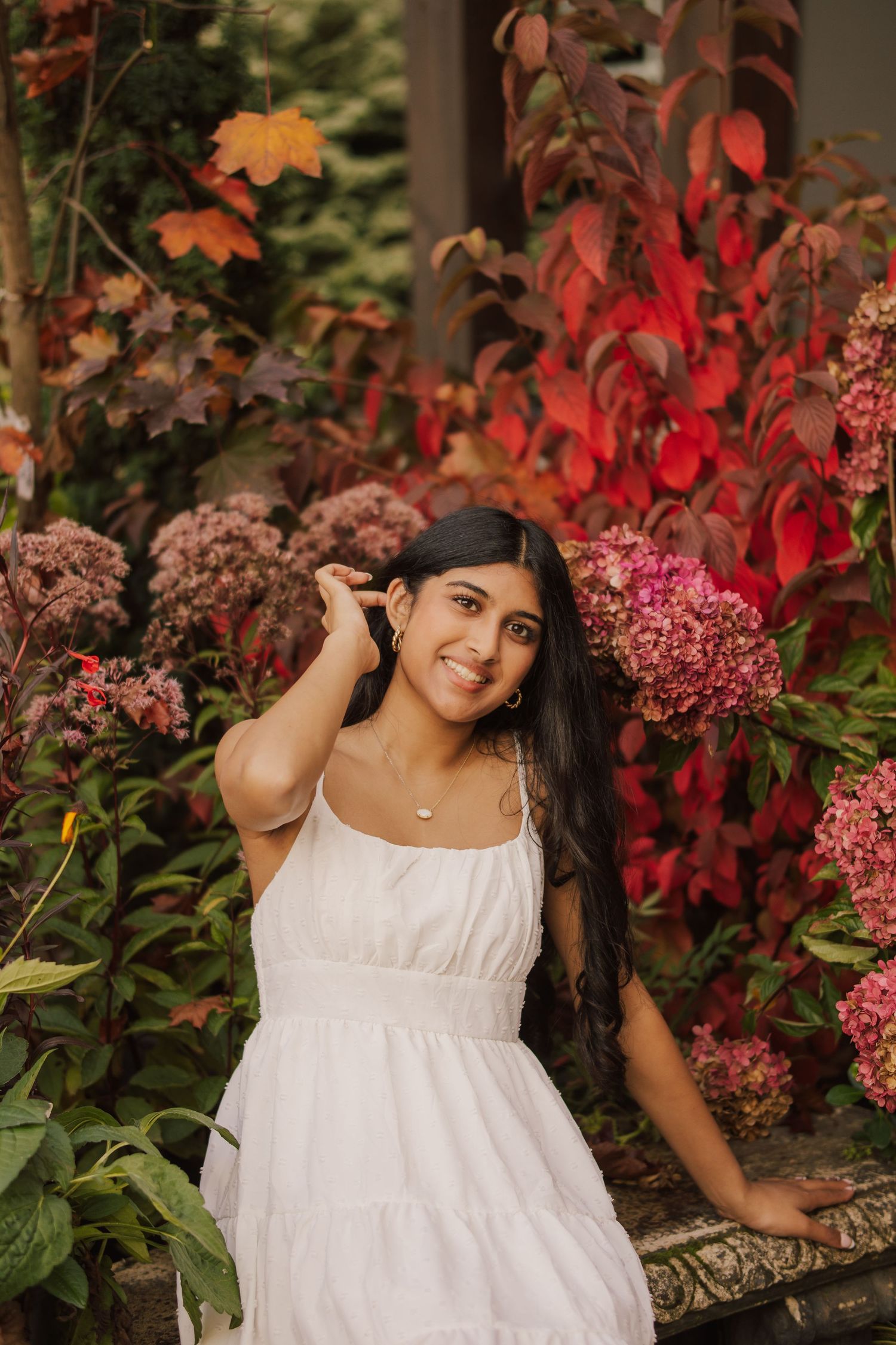 Smiling young woman in a white sundress poses among pink hydrangeas and red autumn foliage in a lush garden.