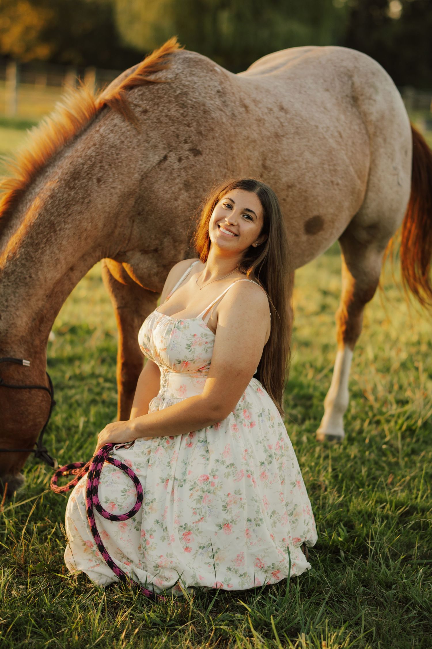 Smiling woman in a floral sundress kneels on green grass holding a pink lead rope beside a grazing roan horse at sunset.