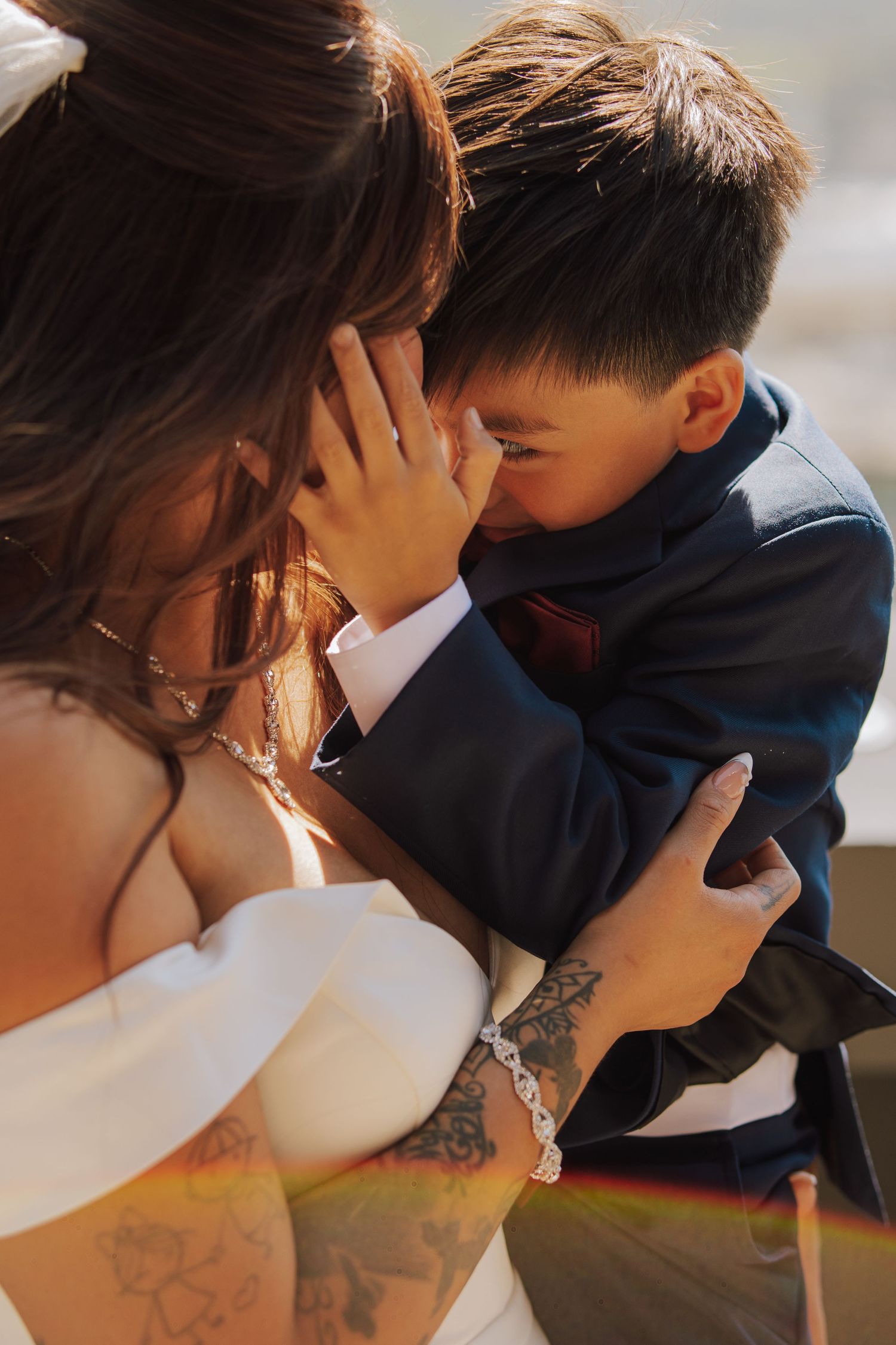 Bride with tattoos embracing an emotional young boy in a navy suit at the wedding.